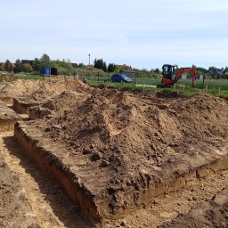 ZBART ELECTRIC Krzysztof Frysztak - Wykopane fundamenty pod budynek z widocznym małym koparką i pracownikami w tle. Wykop ma regularny kształt, ziemia jest jasnobrązowa, a niebo jasne.