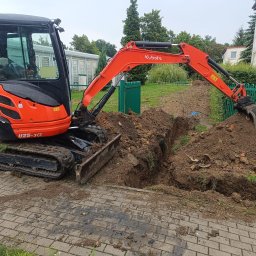 ZBART ELECTRIC Krzysztof Frysztak - Pomarańczowa koparka Kubota U25-3α kopie rów w ziemi obok chodnika z kostki, zielonego płotu i budynku. W tle widoczne drzewa i pochmurne niebo.