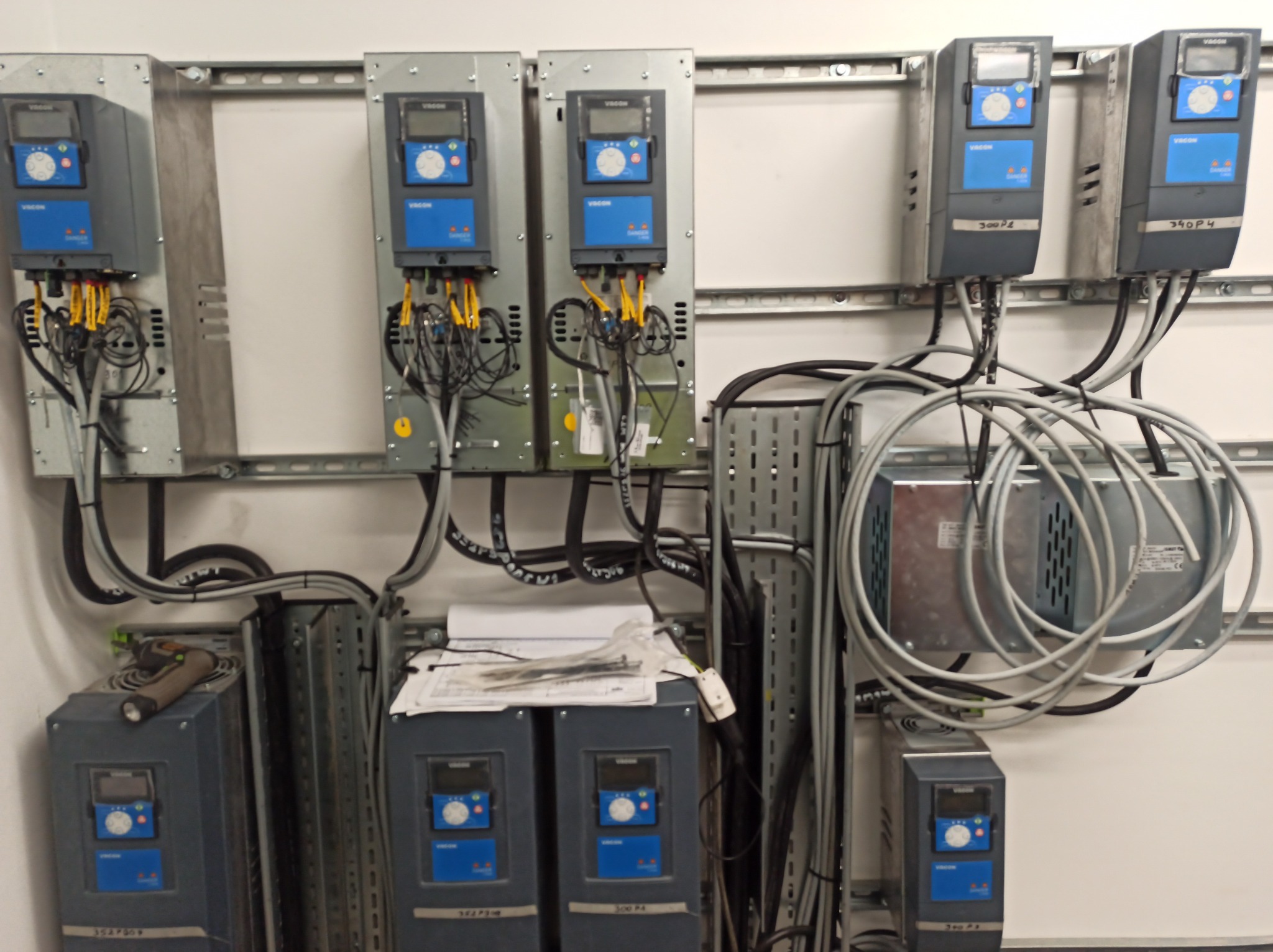 Szafa sterownicza z zamontowanymi falownikami Vacon, okablowaniem i dokumentacją techniczną na białej ścianie. Widoczne narzędzie pracy.