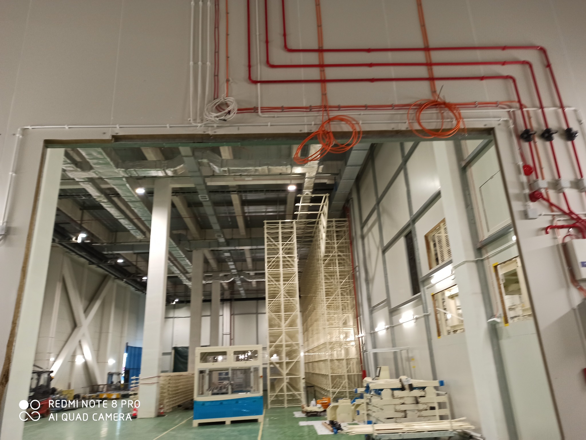 Wnętrze hali produkcyjnej w trakcie budowy, widoczne instalacje elektryczne i rury pod sufitem, regały magazynowe oraz maszyny. Jasne oświetlenie, surowy stan wykończenia.