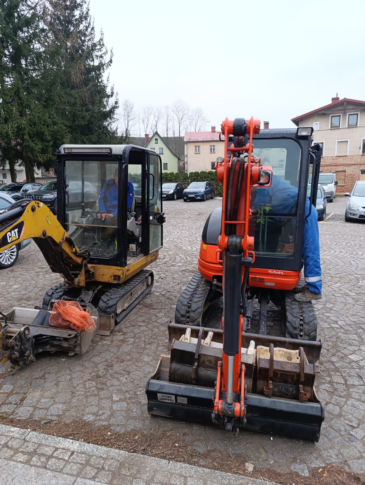 Dwa mini-koparki: żółta CAT z pomarańczową siatką w łyżce i pomarańczowa Kubota z kilkoma łyżkami. Operatorzy w środku. Tło: budynki i zaparkowane samochody.