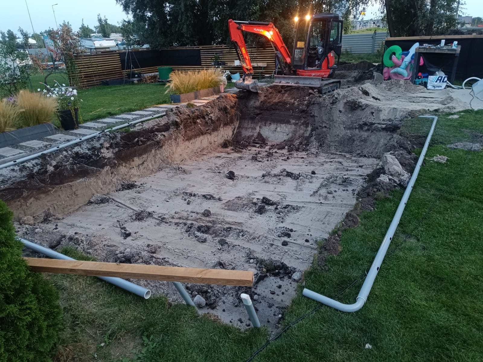 Wykop pod basen w ogrodzie z widocznym minikoparką Kubota i ułożonymi rurami. Etap budowy z ziemią i piaskiem. Elementy dekoracyjne ogrodu w tle.