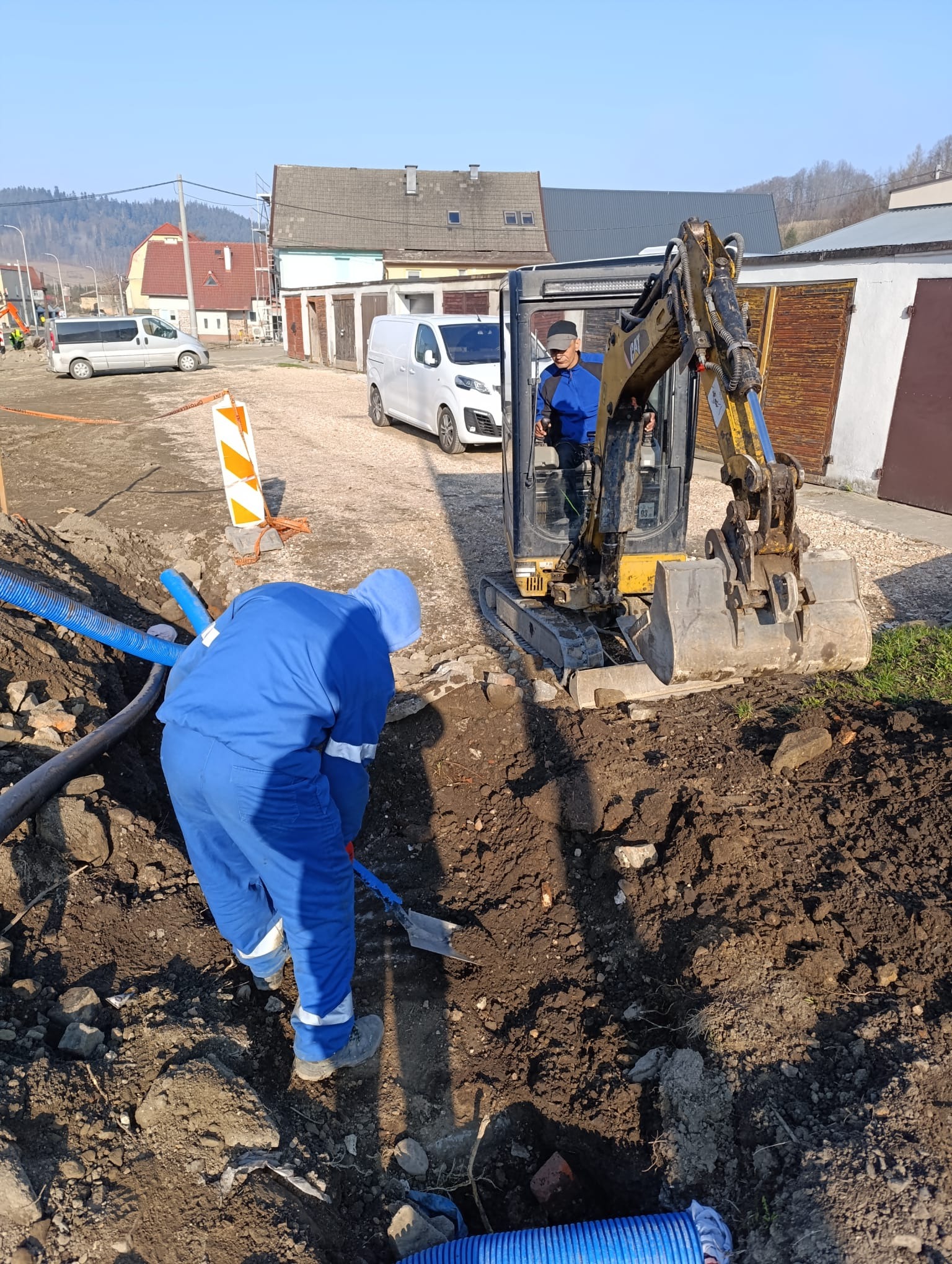 Prace ziemne: operator koparki i pracownik z łopatą przy wykopie z niebieskimi rurami. W tle budynki i pojazdy na tle błękitnego nieba.