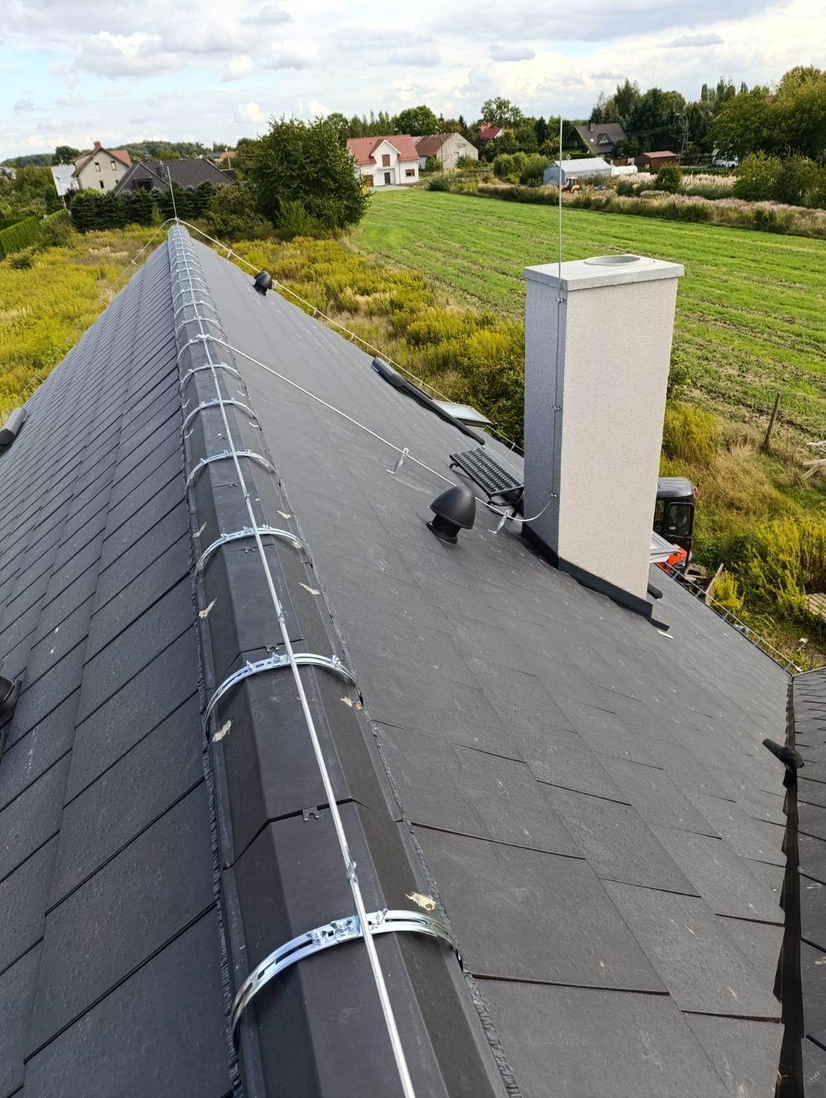 Instalacja odgromowa na dachu z czarnej dachówki, widok z góry. Metalowe uchwyty i przewody biegną wzdłuż kalenicy. W tle domy i zielone pola.