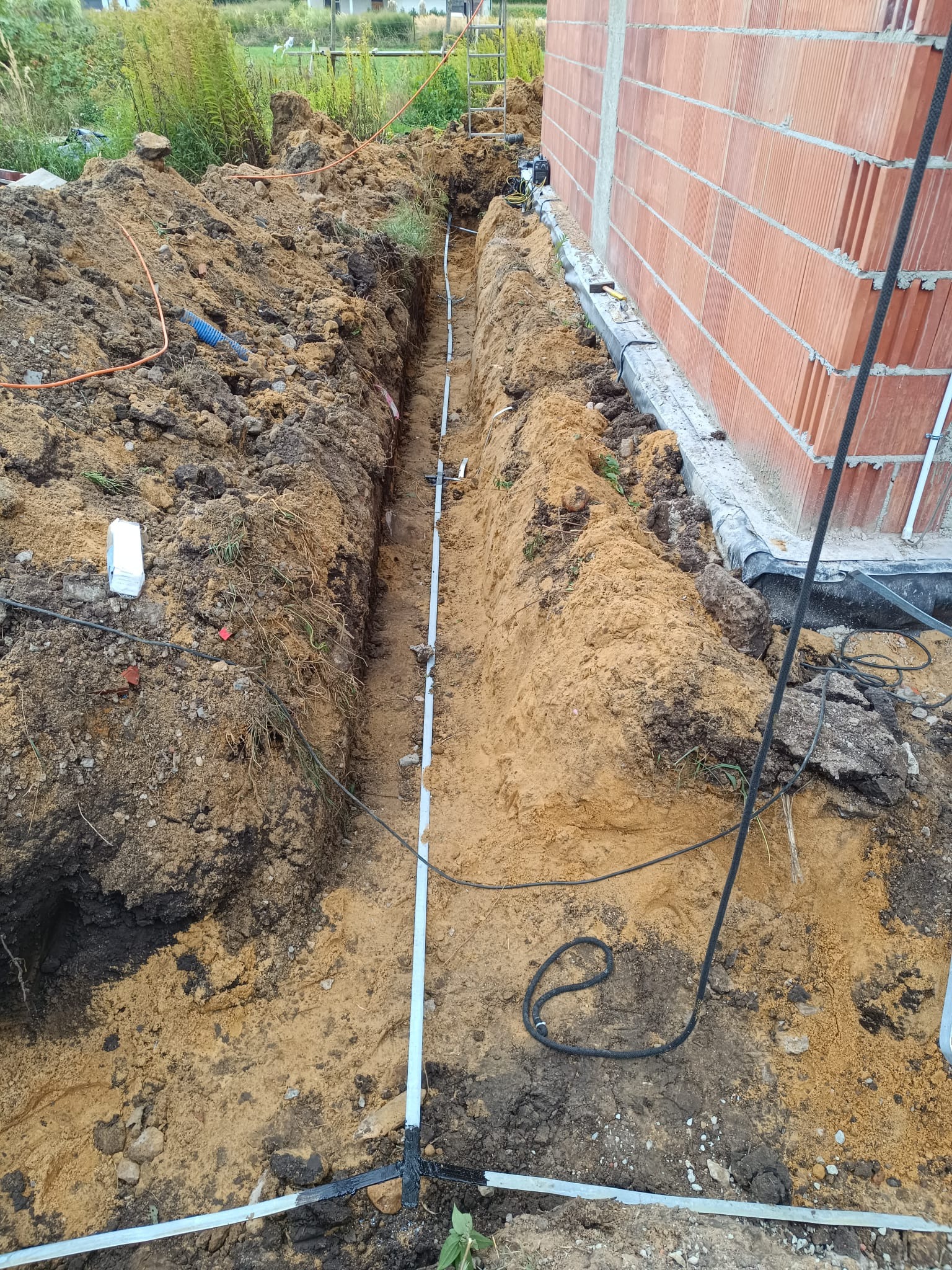 Wykop z ułożonymi rurami osłonowymi kabli elektrycznych w ziemi przy fundamencie domu z czerwonej cegły, widok z góry.