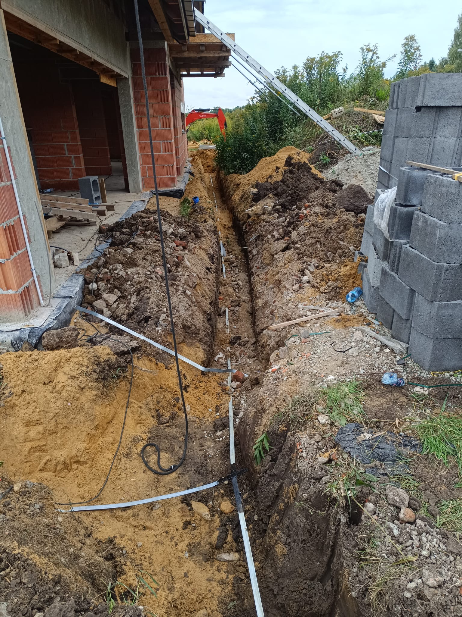 Wykop z rurami instalacyjnymi w ziemi, widok z góry. W tle ceglany budynek w trakcie budowy, drabina i paleta betonowych bloczków.