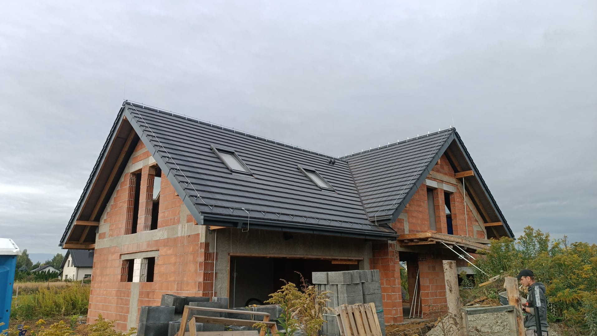 Dom w trakcie budowy: ceglana konstrukcja, czarny dach z oknami dachowymi, widoczne elementy instalacji odgromowej i rusztowania. Szary dzień.