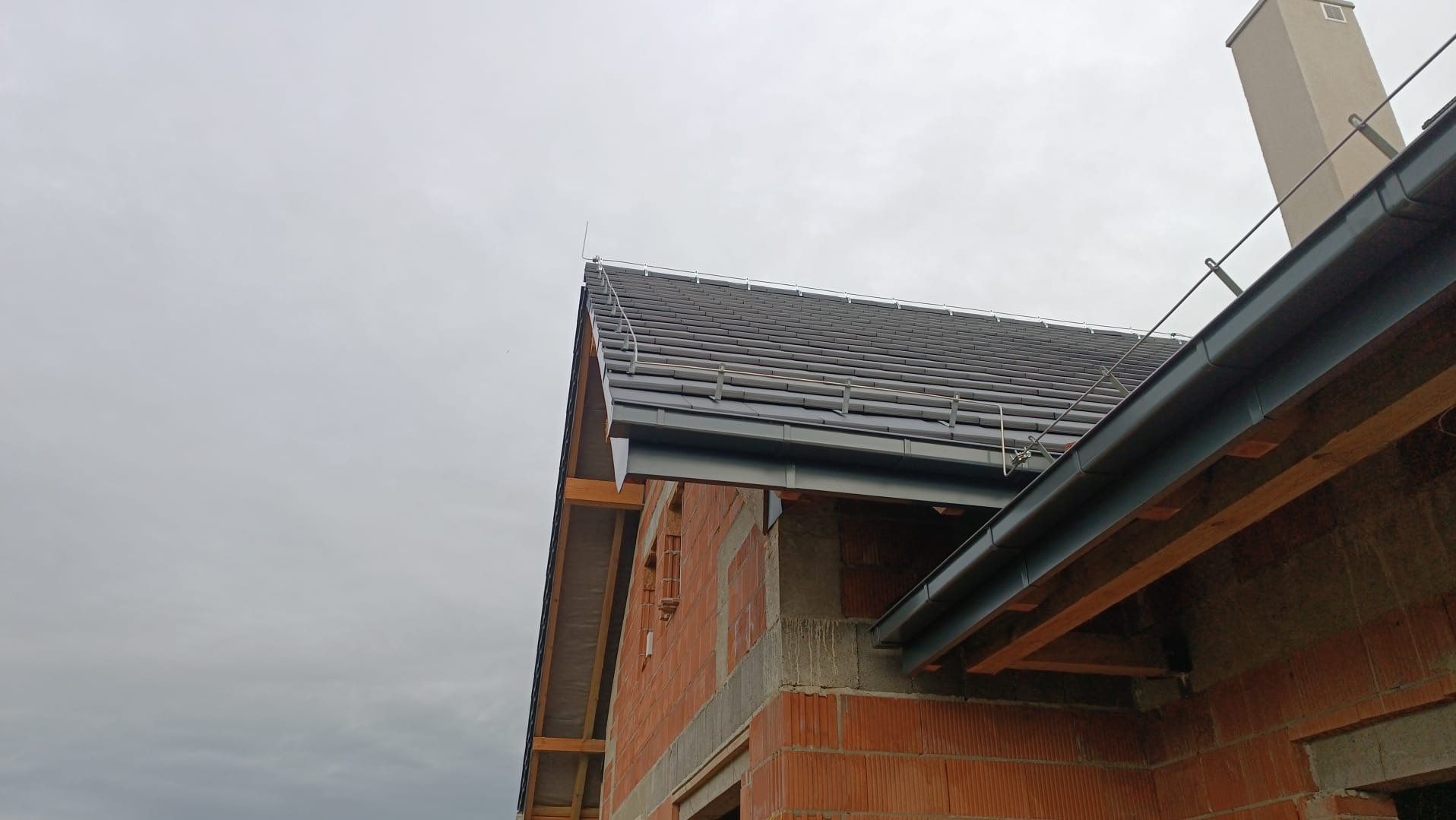 Szary dach z zamontowaną instalacją odgromową na ceglanym budynku w trakcie budowy, widok z dołu pod pochmurnym niebem.