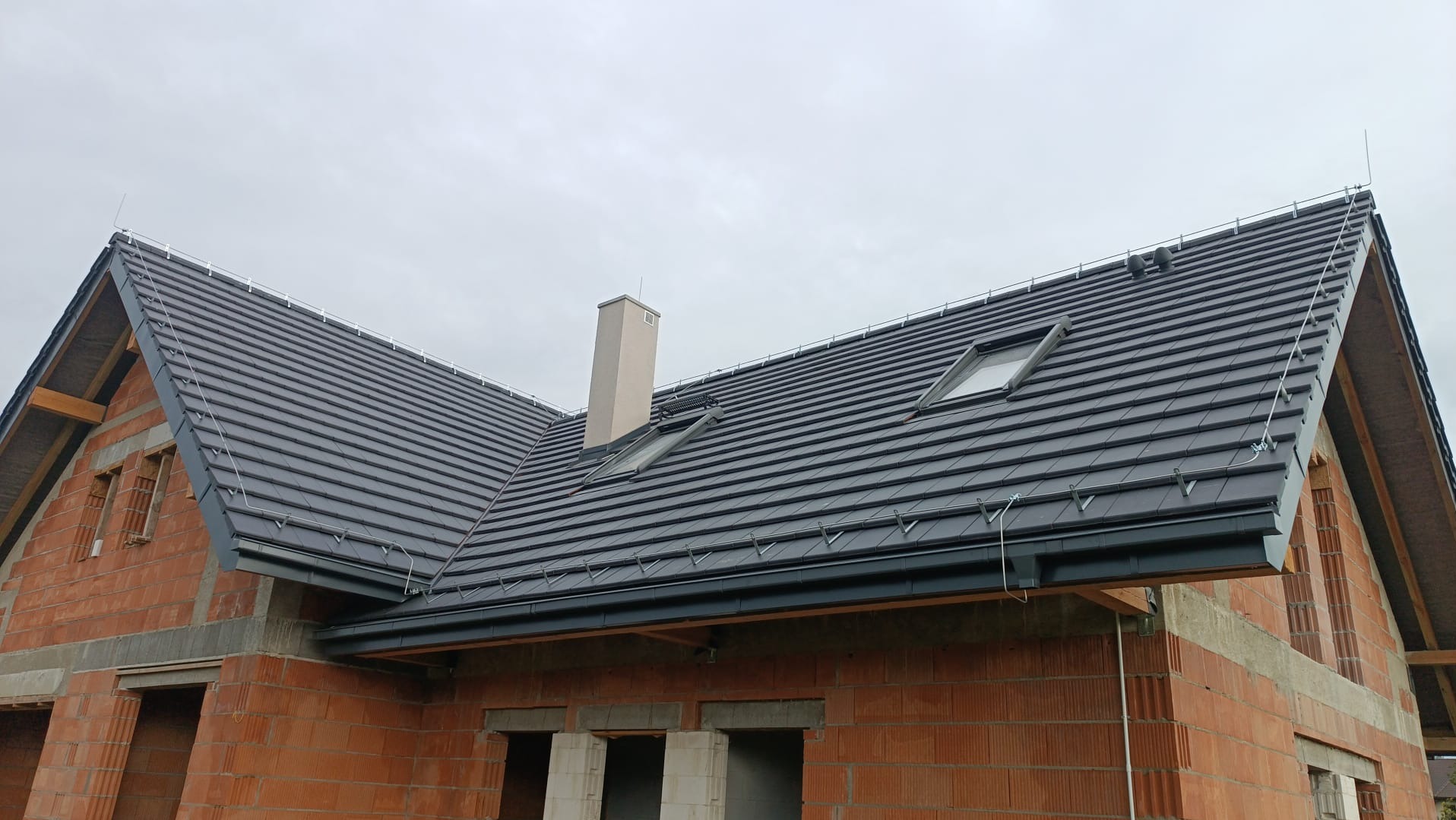 Dom w trakcie budowy z zamontowaną, grafitową dachówką, oknami dachowymi i systemem odgromowym. Widoczne cegły i elementy konstrukcyjne.