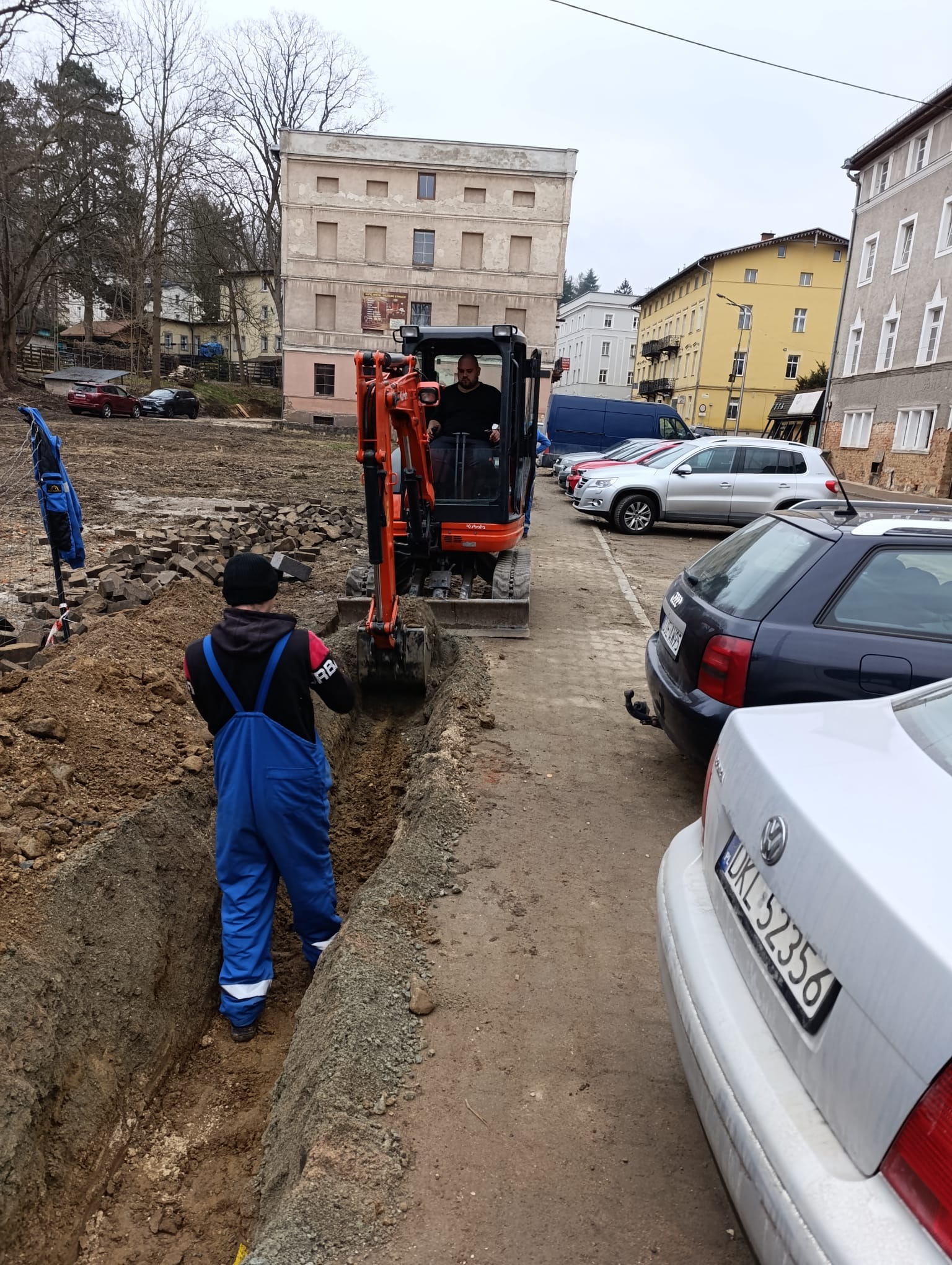 Wykonawca koparką wykopuje rów w ziemi. W tle budynki mieszkalne i zaparkowane auta. Widoczny pracownik w roboczym kombinezonie.