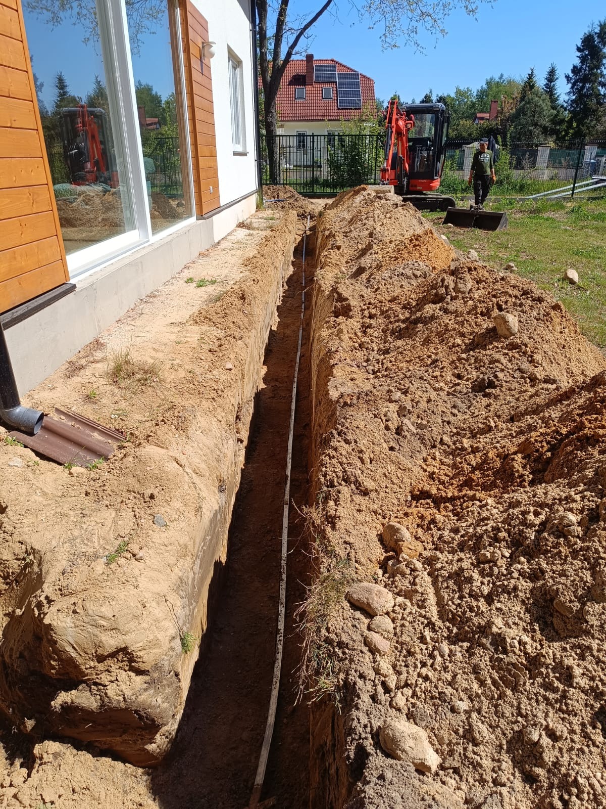 Wykopany rów wzdłuż domu z widoczną rurą, minikoparka i pracownik w tle. Przygotowanie do instalacji podziemnej.