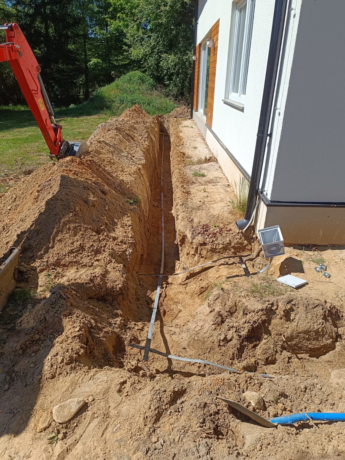Wykop pod instalację elektryczną przy białym budynku jednorodzinnym. W wykopie widoczne rury i przewody. W tle koparka.