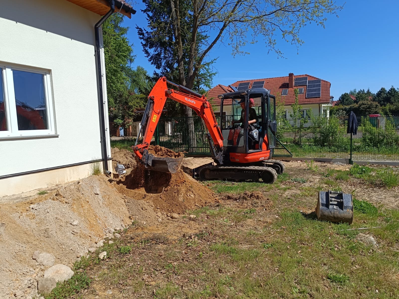 Pomarańczowa minikoparka Kubota w trakcie prac ziemnych przy budynku jednorodzinnym. Widoczny operator i wykopany grunt. W tle dom z panelami słonecznymi.