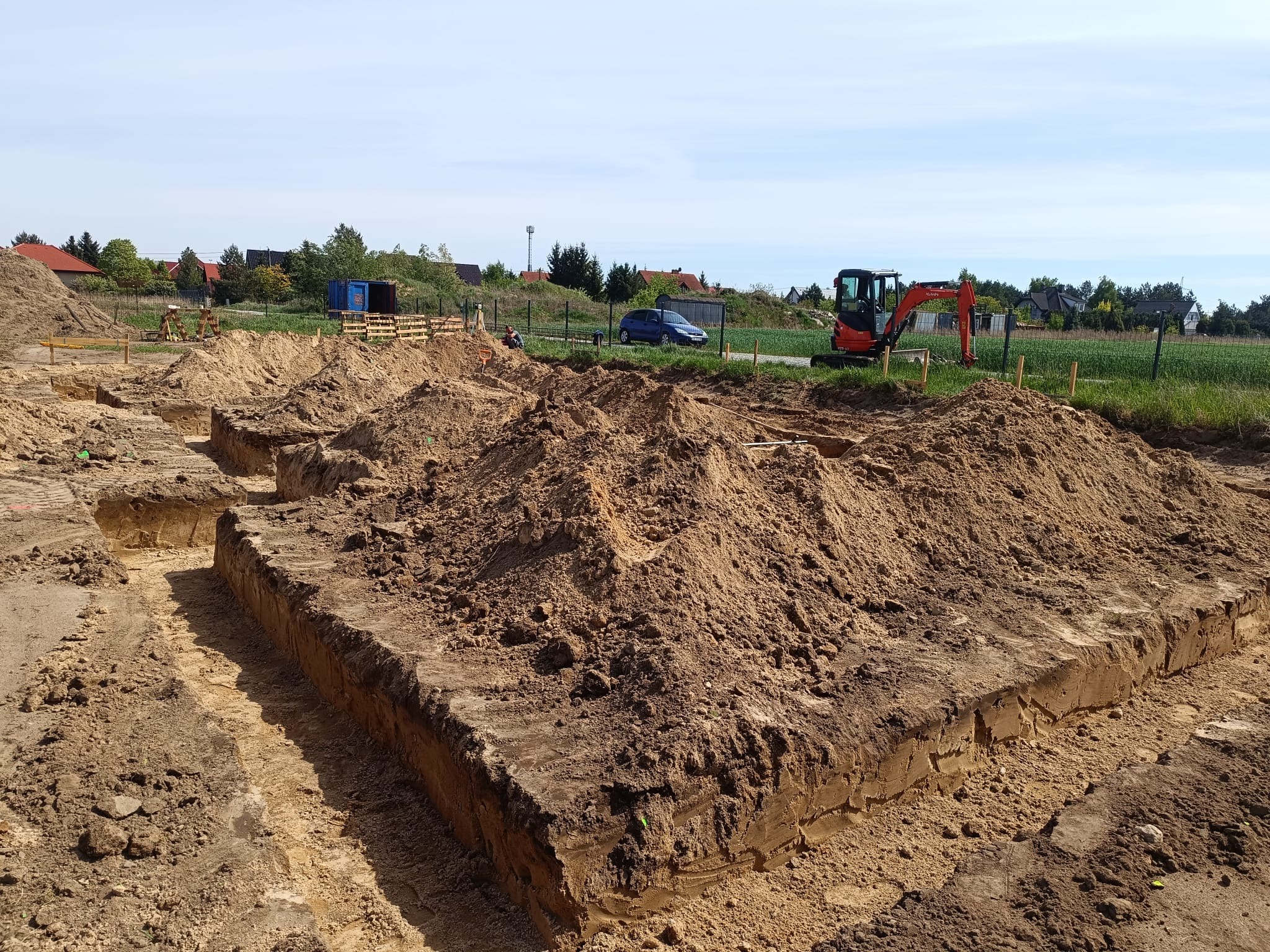 Wykopane fundamenty pod budynek z widocznym małym koparką i pracownikami w tle. Wykop ma regularny kształt, ziemia jest jasnobrązowa, a niebo jasne.