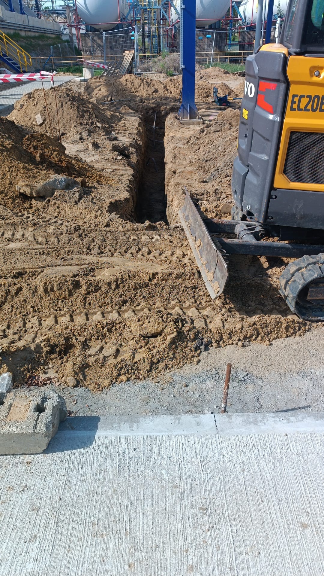 Wykop liniowy w ziemi, obok żółta koparka Volvo EC20E, na tle instalacja przemysłowa i błękitny słup. Widoczne ślady opon i przesuniętej ziemi.