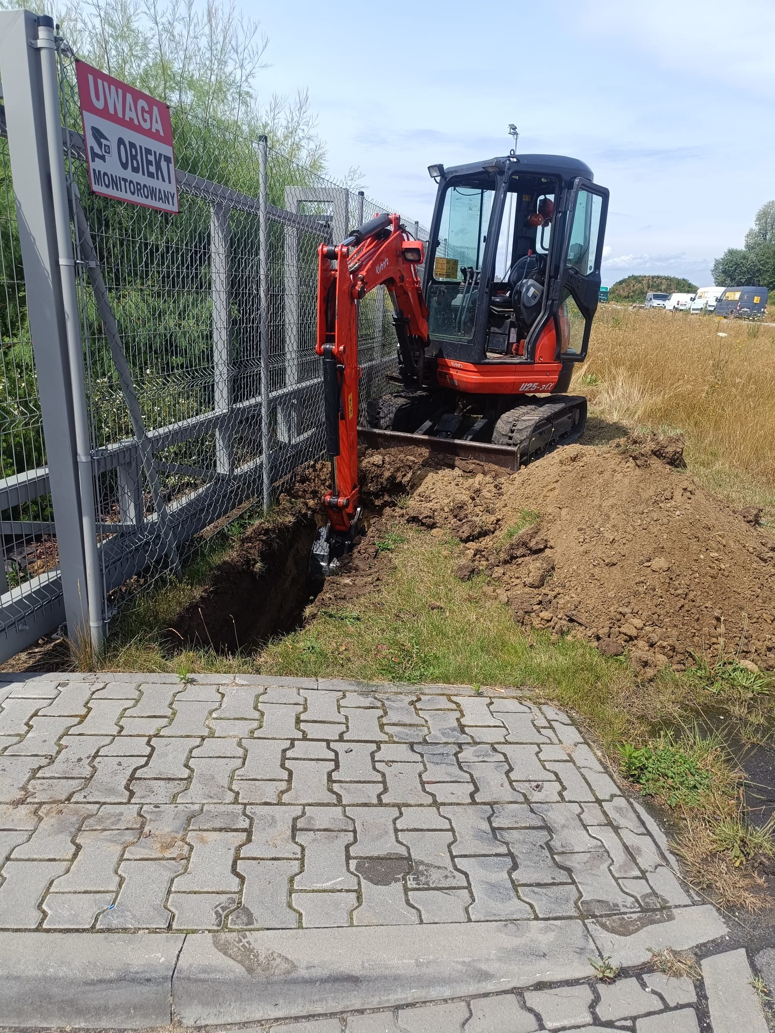 Pomarańczowa mini koparka Kubota U25-3α kopie rów w ziemi obok szarego ogrodzenia z siatki z tabliczką 'Uwaga obiekt monitorowany'.