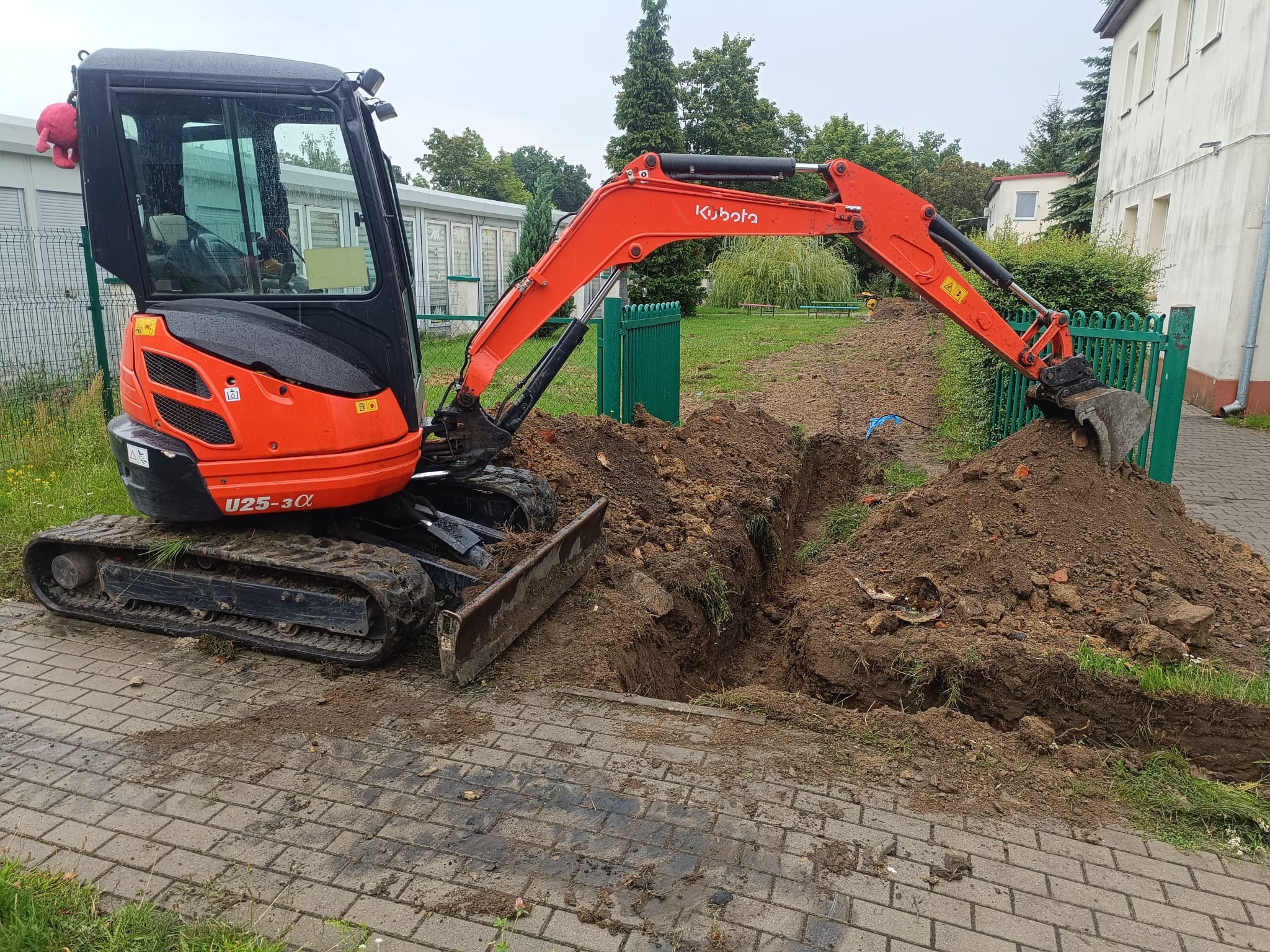 Pomarańczowa koparka Kubota U25-3α kopie rów w ziemi obok chodnika z kostki, zielonego płotu i budynku. W tle widoczne drzewa i pochmurne niebo.
