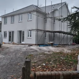 Dwupiętrowy dom z szarą elewacją i dachem, otoczony rusztowaniami. Widoczny fragment samochodu dostawczego i ogród z opadłymi liśćmi. Pochmurne niebo w tle.