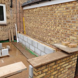 WW Building co Ltd - Murowana ściana z bloczków i cegieł w trakcie budowy, widoczne narzędzia i materiały budowlane, z żółtą cegłą w tle. Detal prac murarskich.