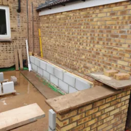 Murowana ściana z bloczków i cegieł w trakcie budowy, widoczne narzędzia i materiały budowlane, z żółtą cegłą w tle. Detal prac murarskich.