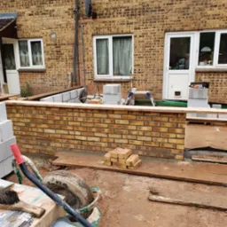 Budowa niskiego murku z cegły klinkierowej przy budynku mieszkalnym. Widoczne narzędzia budowlane i materiały przygotowane do dalszych prac murarskich.