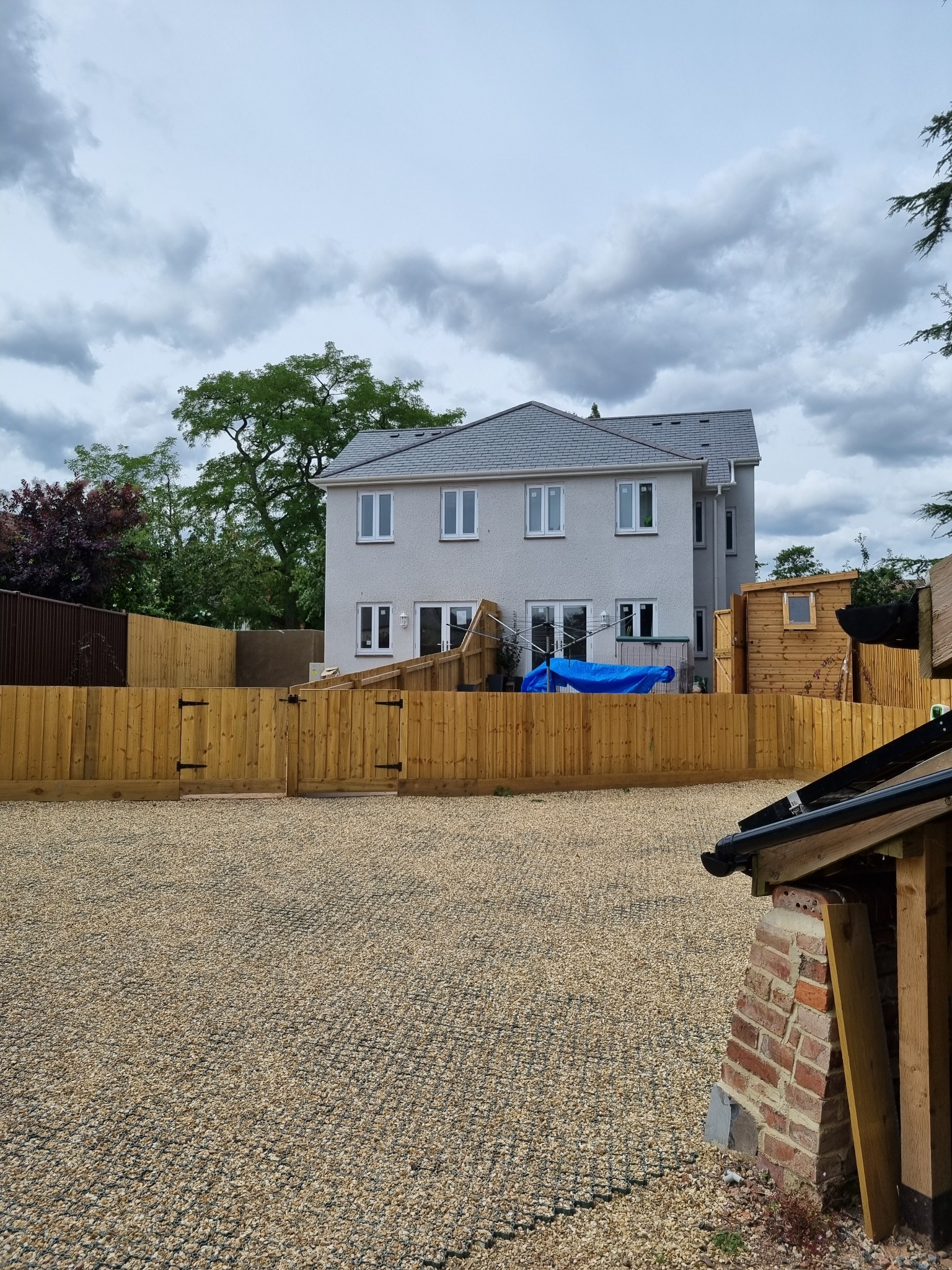 Dwupiętrowy dom z szarą dachówką, otoczony drewnianym ogrodzeniem i żwirowym podłożem. Widok na elewację z białymi oknami. Na podwórku widoczny niebieski materiał.