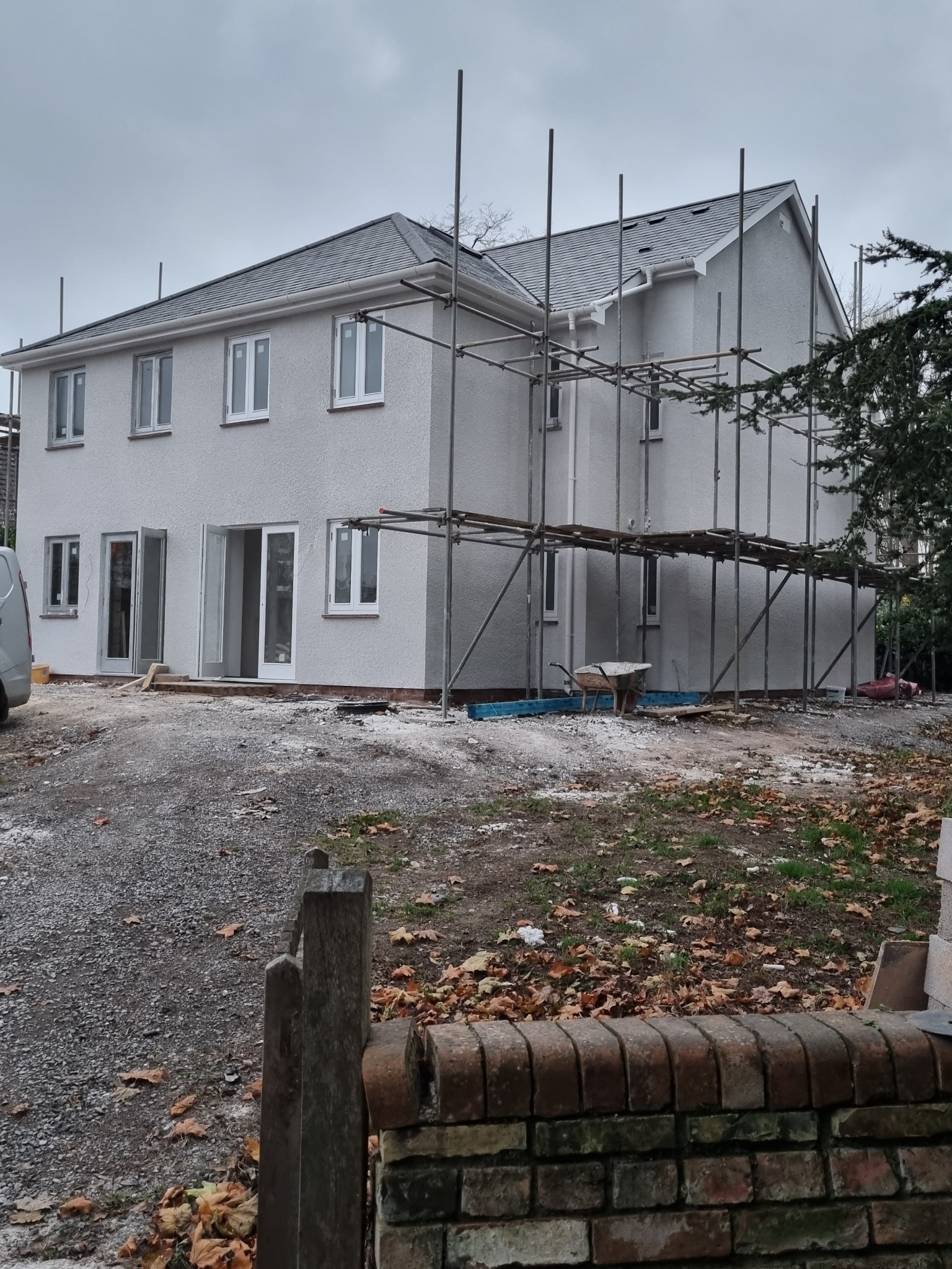 Dwupiętrowy dom z szarą elewacją i dachem, otoczony rusztowaniami. Widoczny fragment samochodu dostawczego i ogród z opadłymi liśćmi. Pochmurne niebo w tle.