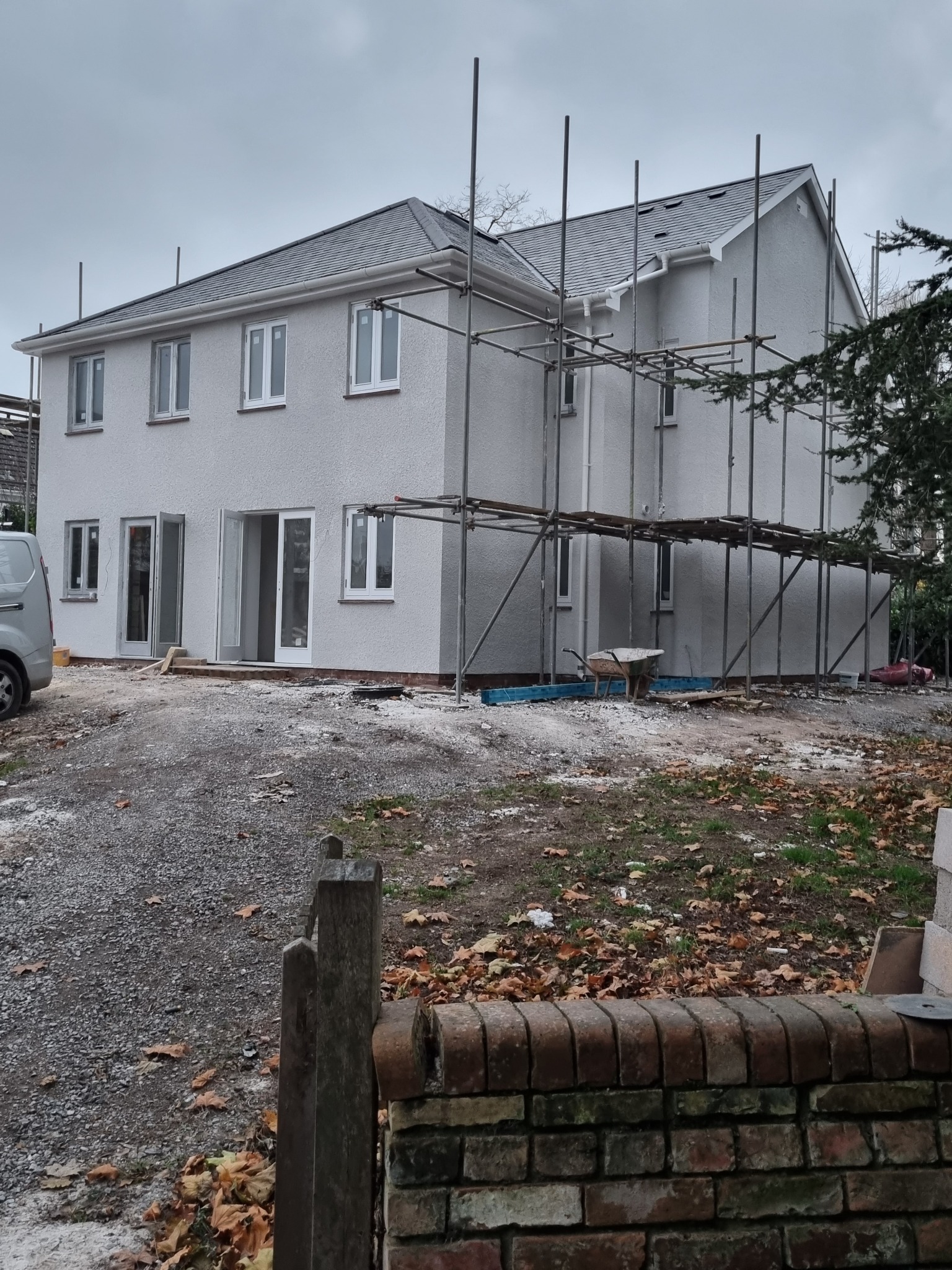 Dwupiętrowy dom z szarą elewacją i białymi oknami, otoczony rusztowaniem. Widoczny fragment samochodu dostawczego i ceglany murek z drewnianym słupkiem na pierwszym planie.
