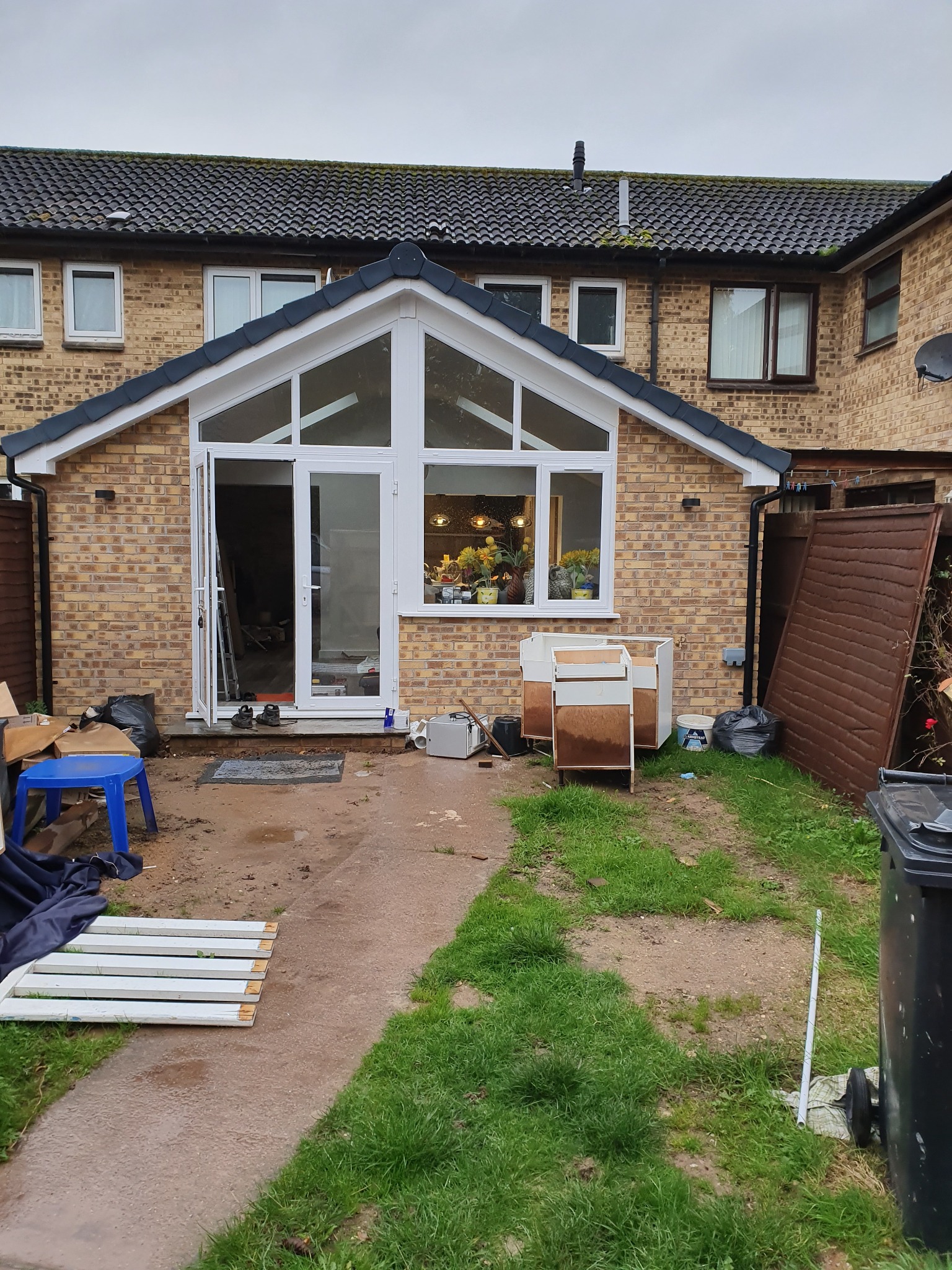 Rozbudowa domu z cegły z dużymi oknami i otwartymi drzwiami. Widoczne materiały budowlane i narzędzia sugerują trwające prace wykończeniowe.