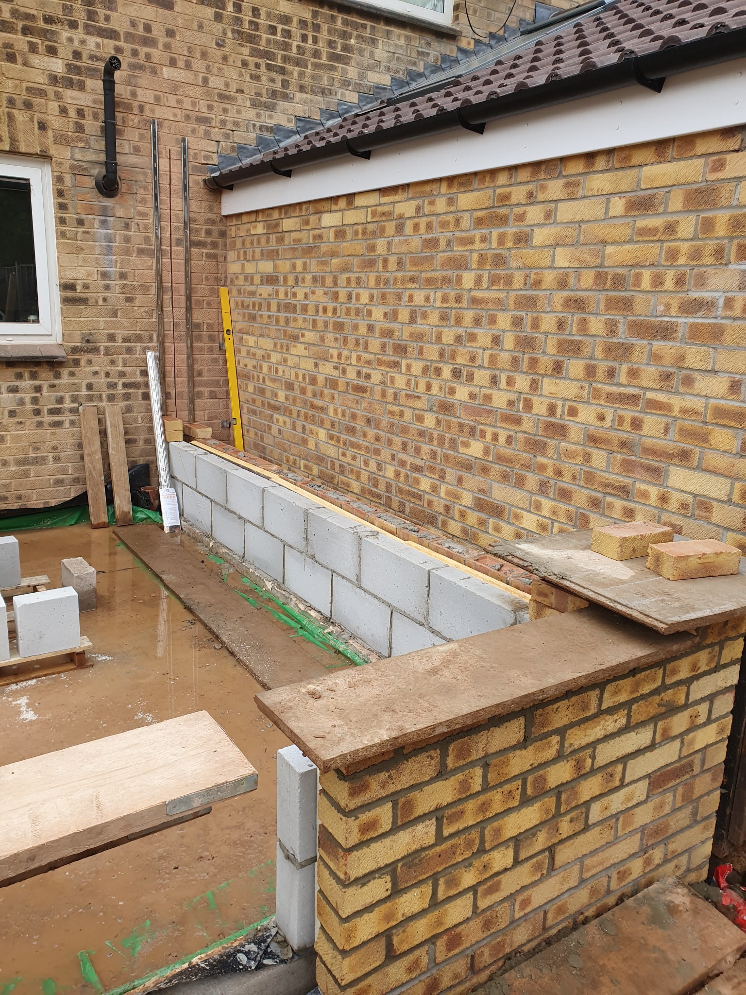 Murowana ściana z bloczków i cegieł w trakcie budowy, widoczne narzędzia i materiały budowlane, z żółtą cegłą w tle. Detal prac murarskich.