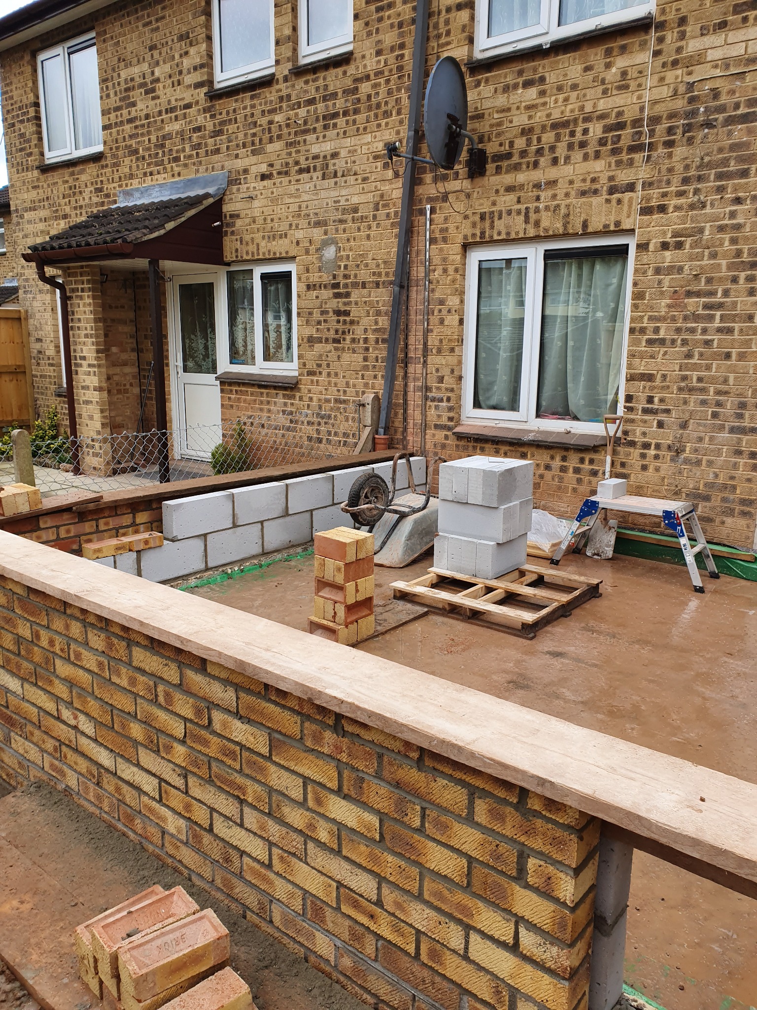 Budowa murku z cegły klinkierowej i bloczków betonowych na posesji, widoczne narzędzia i materiały budowlane. Etap prac murarskich na zewnątrz domu.