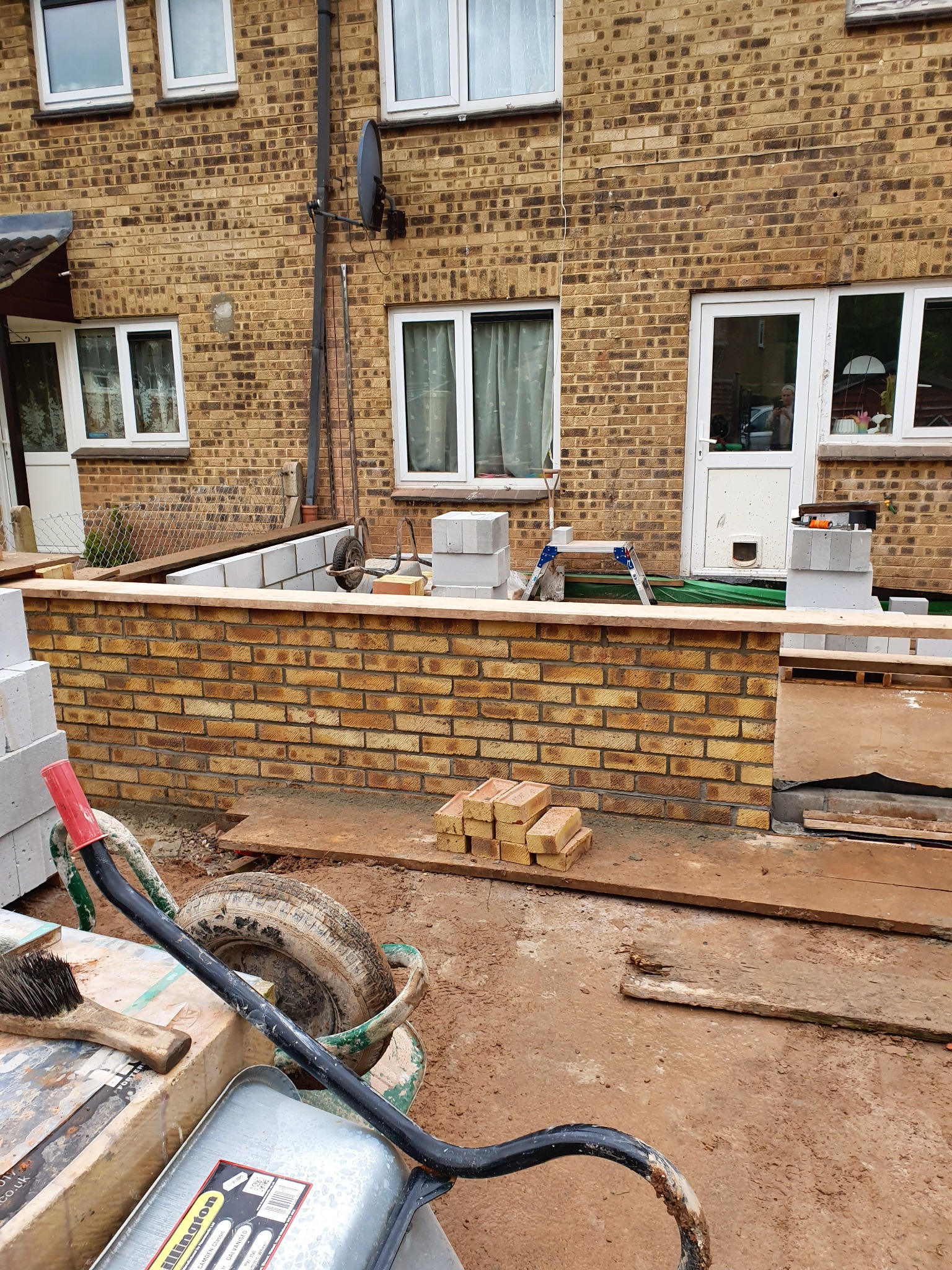 Budowa niskiego murku z cegły klinkierowej przy budynku mieszkalnym. Widoczne narzędzia budowlane i materiały przygotowane do dalszych prac murarskich.