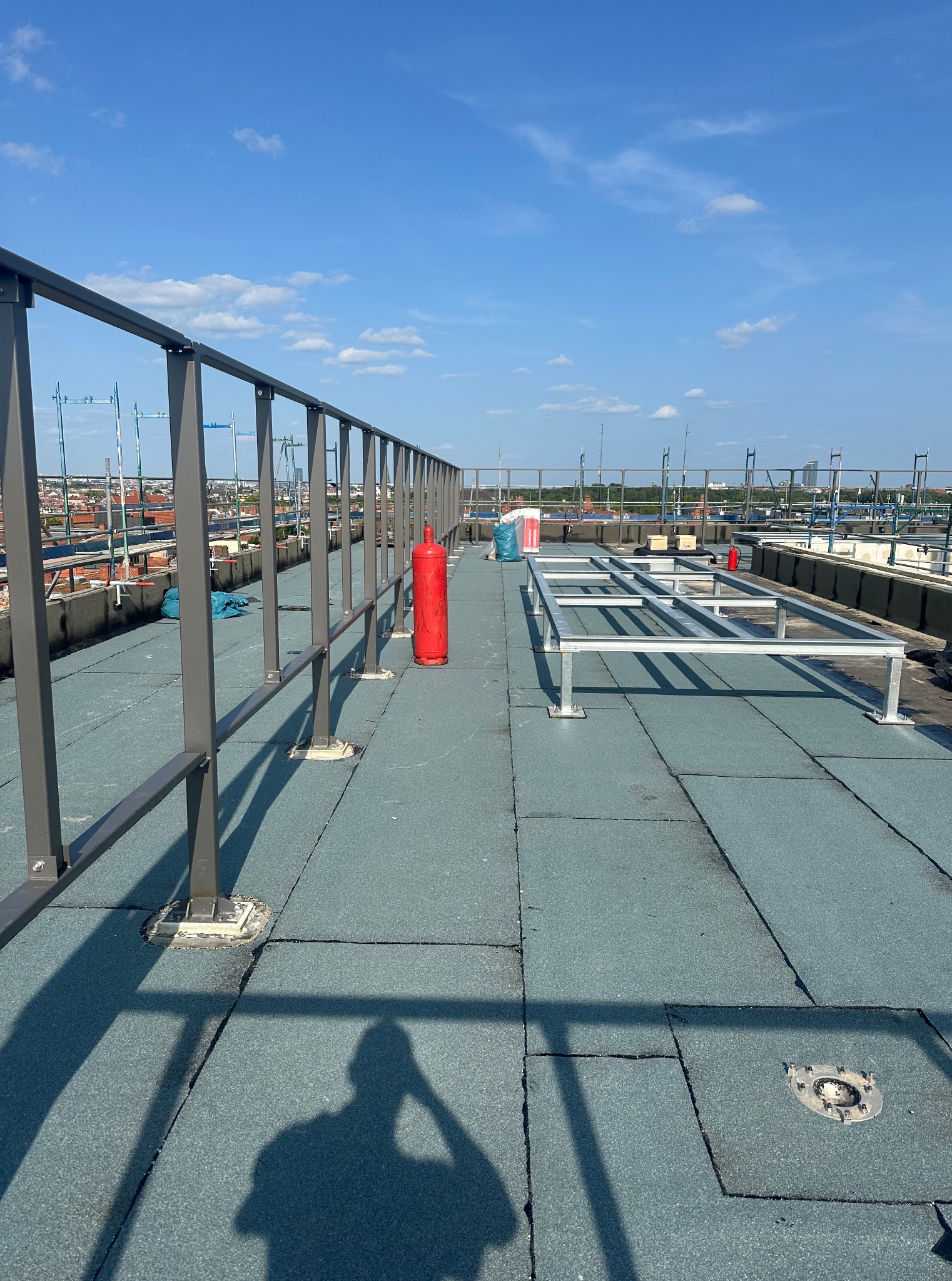 Płaski dach pokryty szarozieloną papą, z metalową barierką i czerwoną butlą. W tle zabudowa miejska pod błękitnym niebem z chmurami. Widoczny cień osoby.