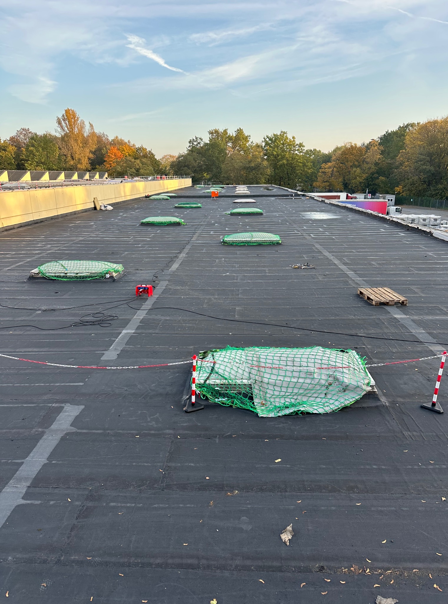 Rozległy, płaski dach pokryty czarną papą, zabezpieczony siatkami i barierkami. Widoczne świetliki dachowe i narzędzia. Praca na wysokości w jesiennym krajobrazie.