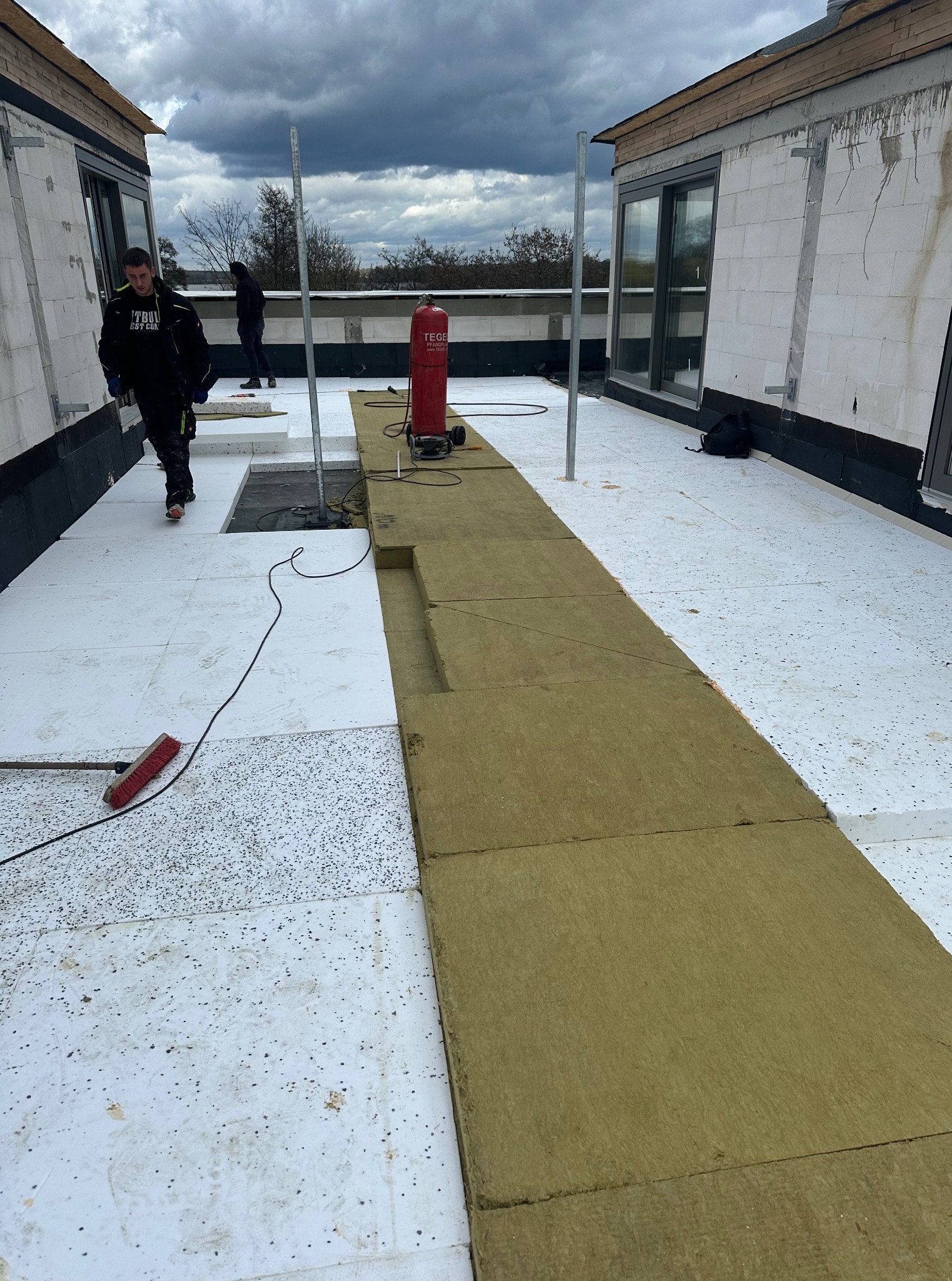 Montaż izolacji termicznej na dachu płaskim z użyciem wełny mineralnej. Widoczny pracownik, narzędzia i elementy konstrukcyjne budynku w trakcie prac dekarskich.