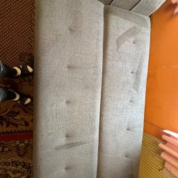 Fresh Space - Szara kanapa z widocznymi plamami na tapicerce, sfotografowana z góry, obok dywan z wzorem i fragment pomarańczowej ściany. Widoczne buty osoby stojącej obok.