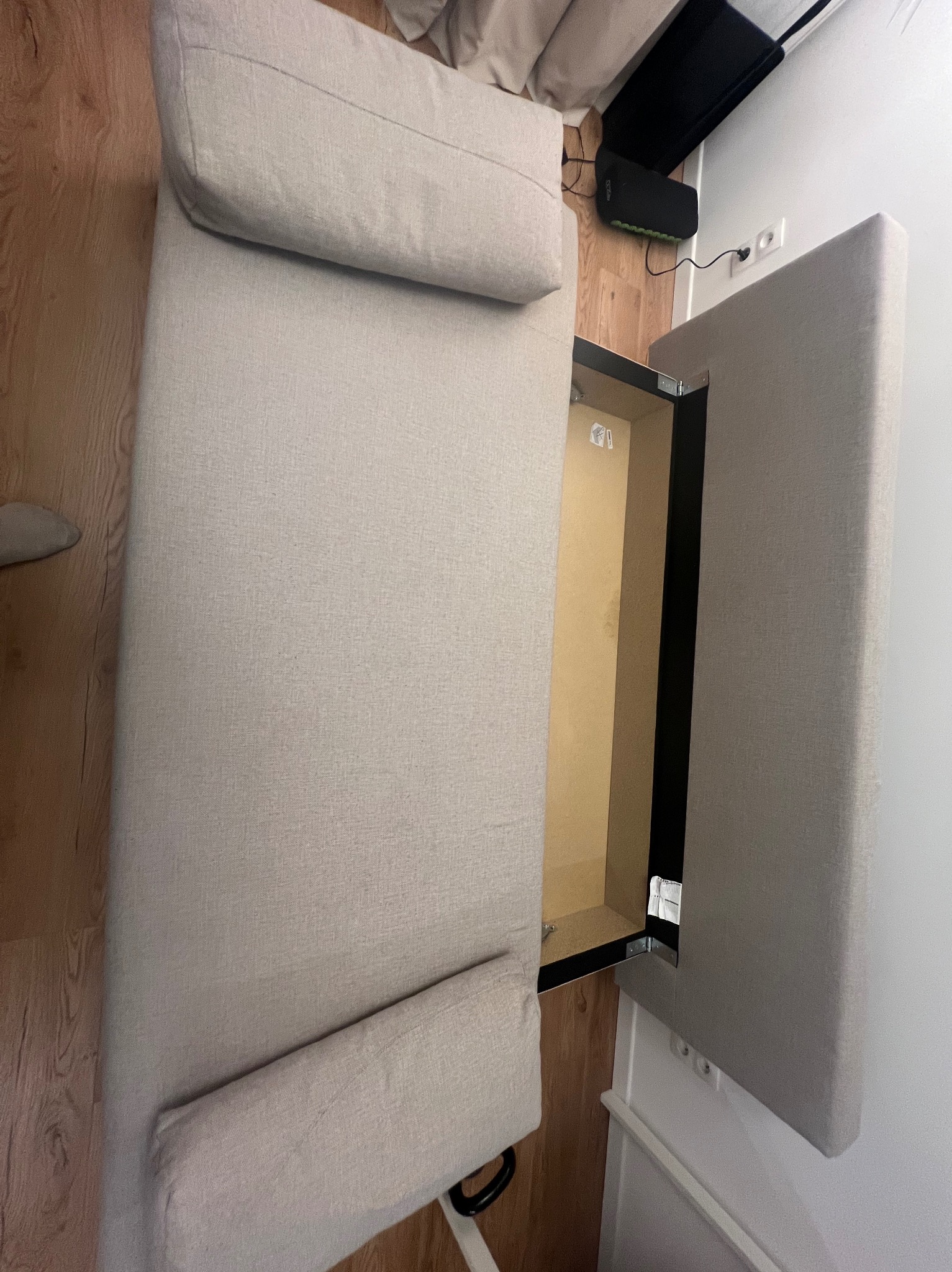 Jasna kanapa z otwieranym pojemnikiem na pościel, widok z góry, ukazujący wnętrze pojemnika. Minimalistyczny design, drewniana podłoga w tle.