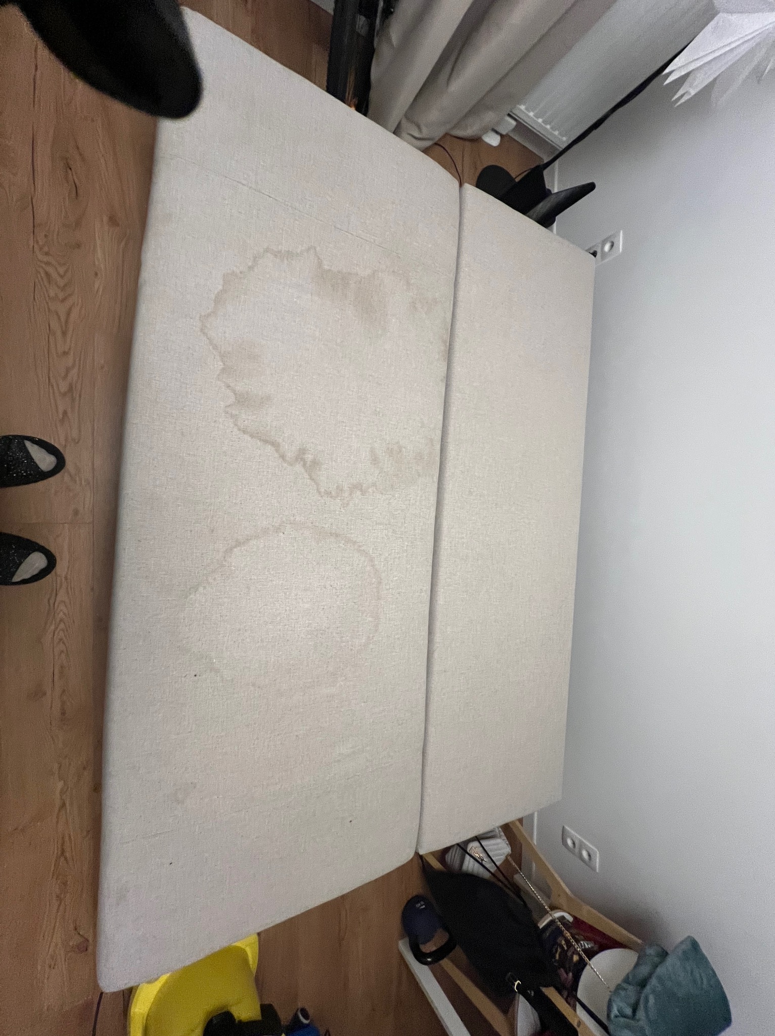 Beżowa kanapa z widocznymi plamami na tapicerce, ustawiona w rogu pokoju. Widoczne drewniane panele podłogowe i biała ściana.