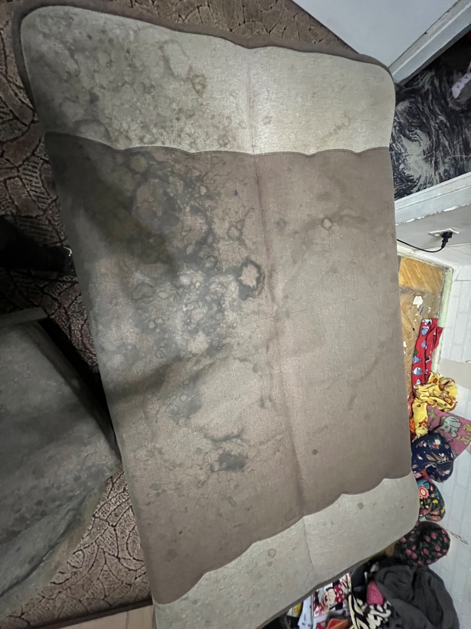 Widok z góry na zabrudzoną kanapę z plamami, stojącą w pokoju z widocznymi ubraniami i wzorzystą tapetą. Potrzeba czyszczenia tapicerki.