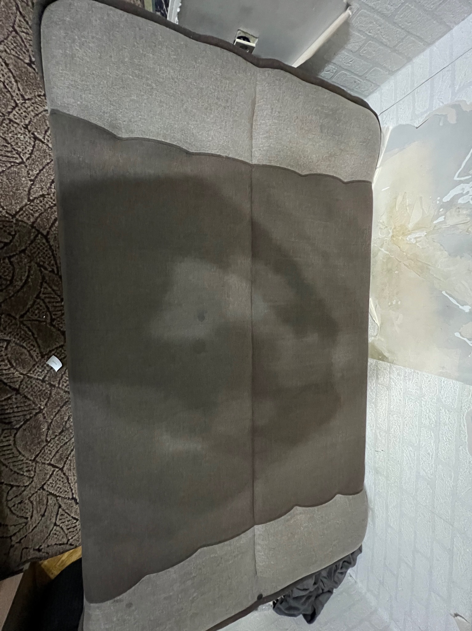 Szary narożnik z widocznymi plamami po czyszczeniu, stojący na tle wzorzystego dywanu i ściany z tapetą imitującą cegłę. Widoczne ślady wilgoci na narożniku.