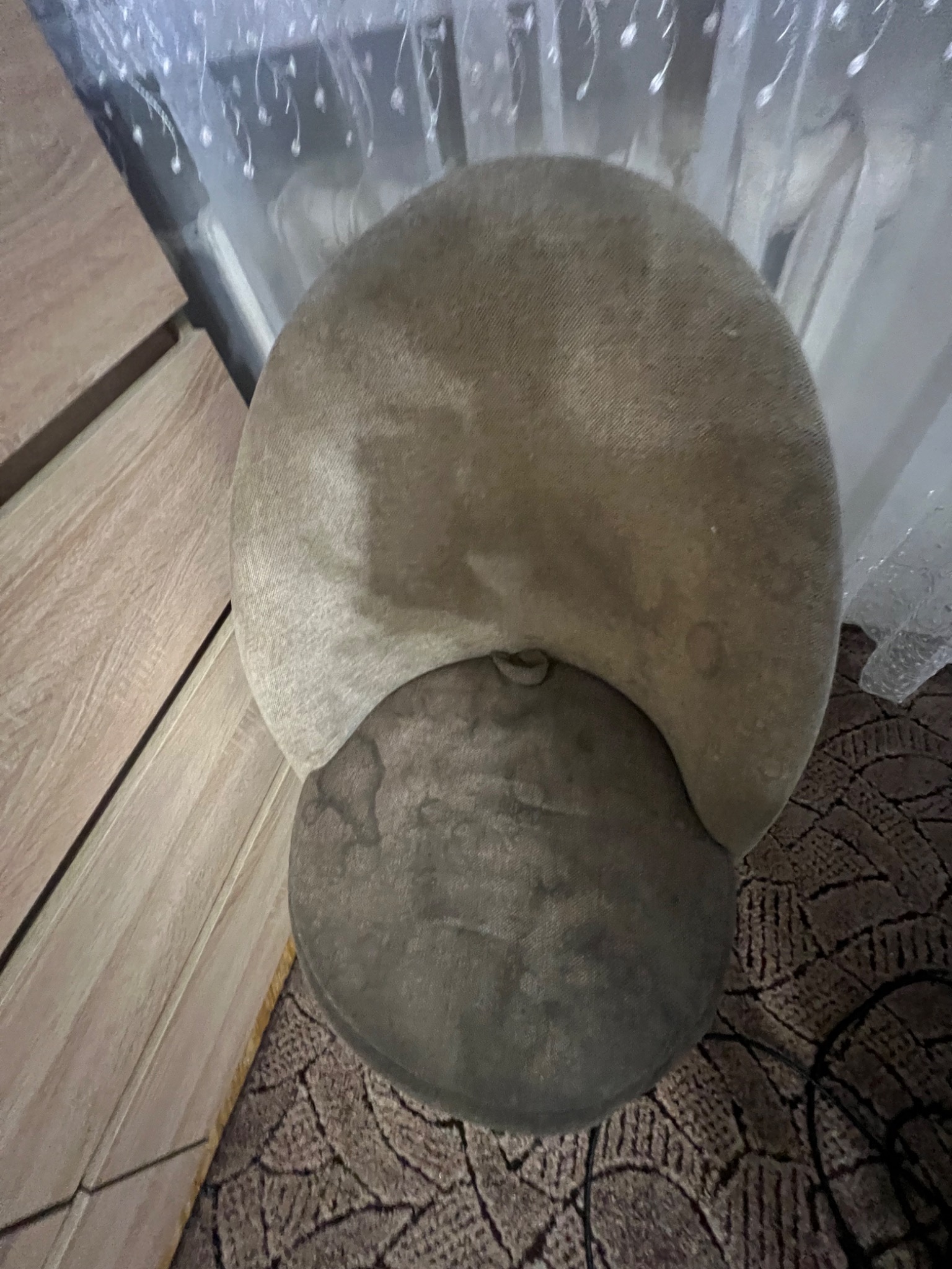 Dwu-segmentowy, okrągły taboret tapicerowany beżową tkaniną, widziany z góry na tle wzorzystego dywanu i mebli. Widoczne plamy na tapicerce.