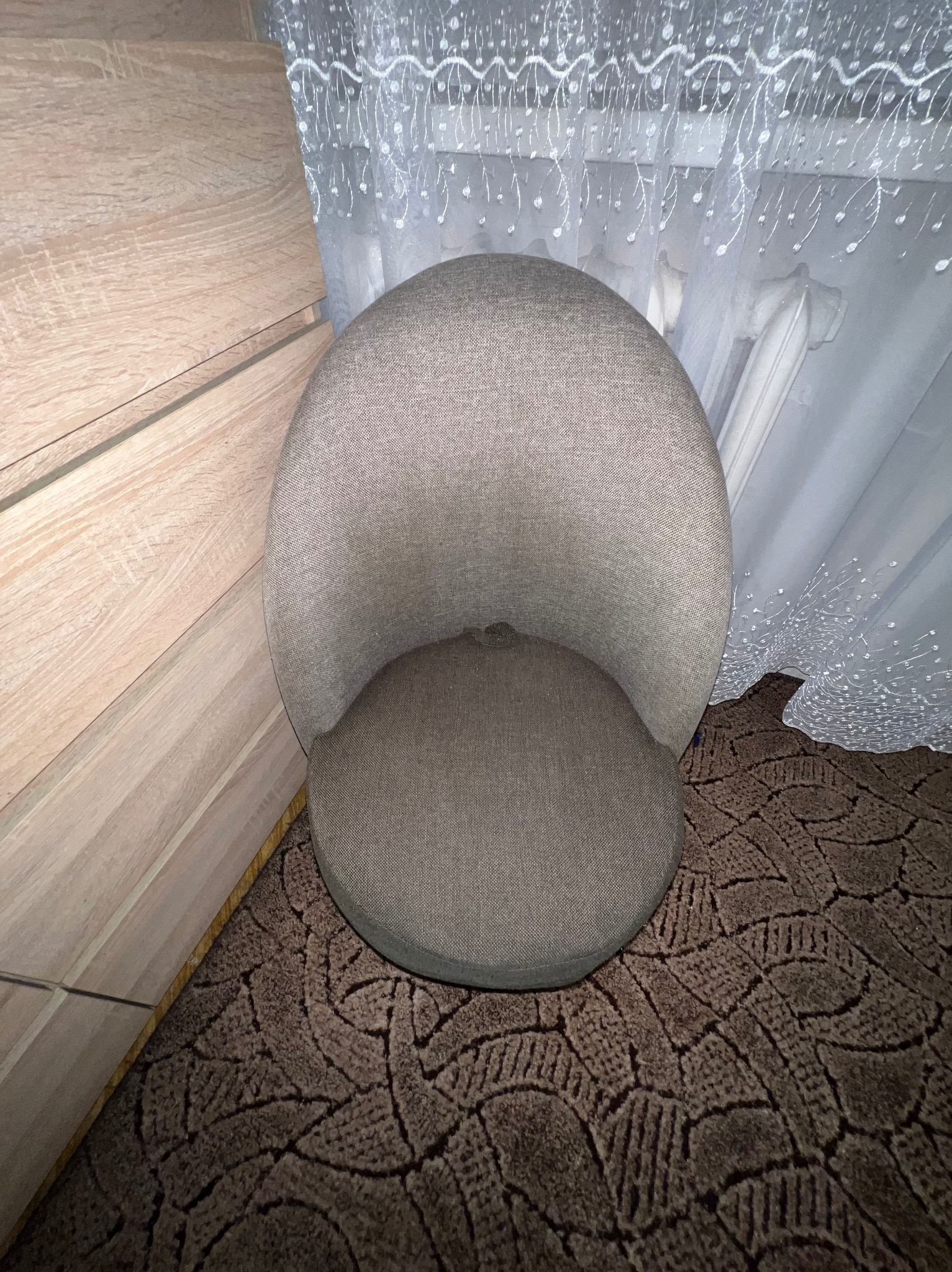 Szary, okrągły fotel tapicerowany stoi na brązowym dywanie z wzorem, obok drewnianej komody i białej, wzorzystej firanki. Widok z góry.