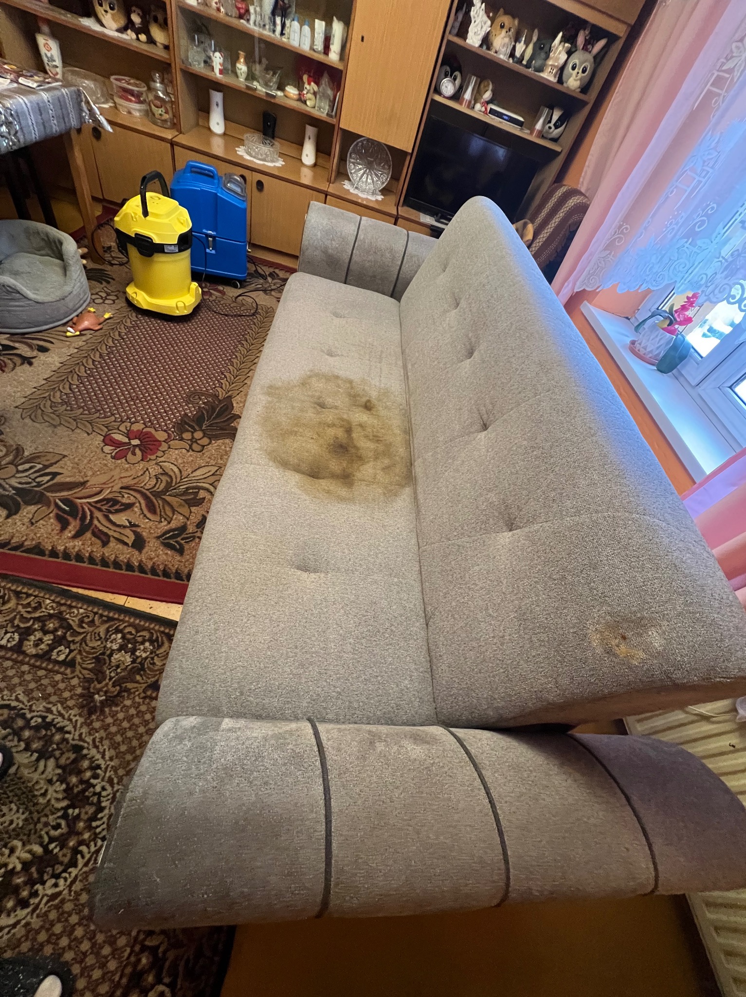 Szara sofa z widoczną plamą na siedzisku, sprzęt do czyszczenia obok. Usługi czyszczenia tapicerki w Gdańsku. Widok z góry, wnętrze mieszkania.
