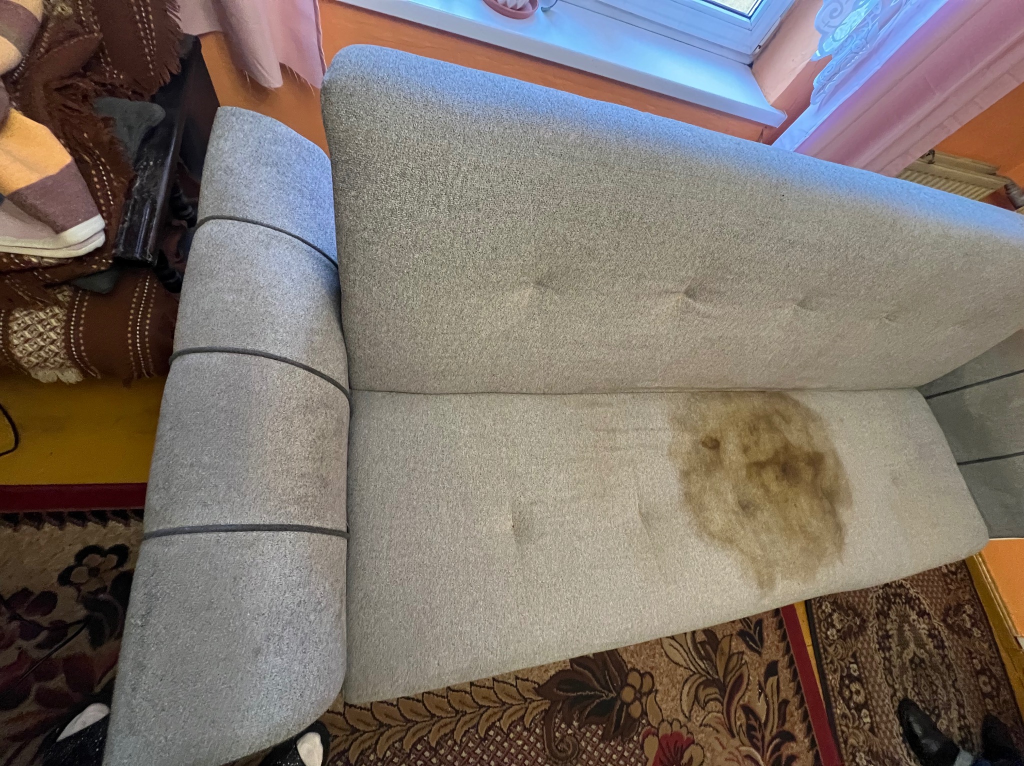 Szara kanapa z widoczną, dużą plamą na siedzisku, ukazująca potrzebę profesjonalnego czyszczenia tapicerki. Widok z góry, dywan w tle.