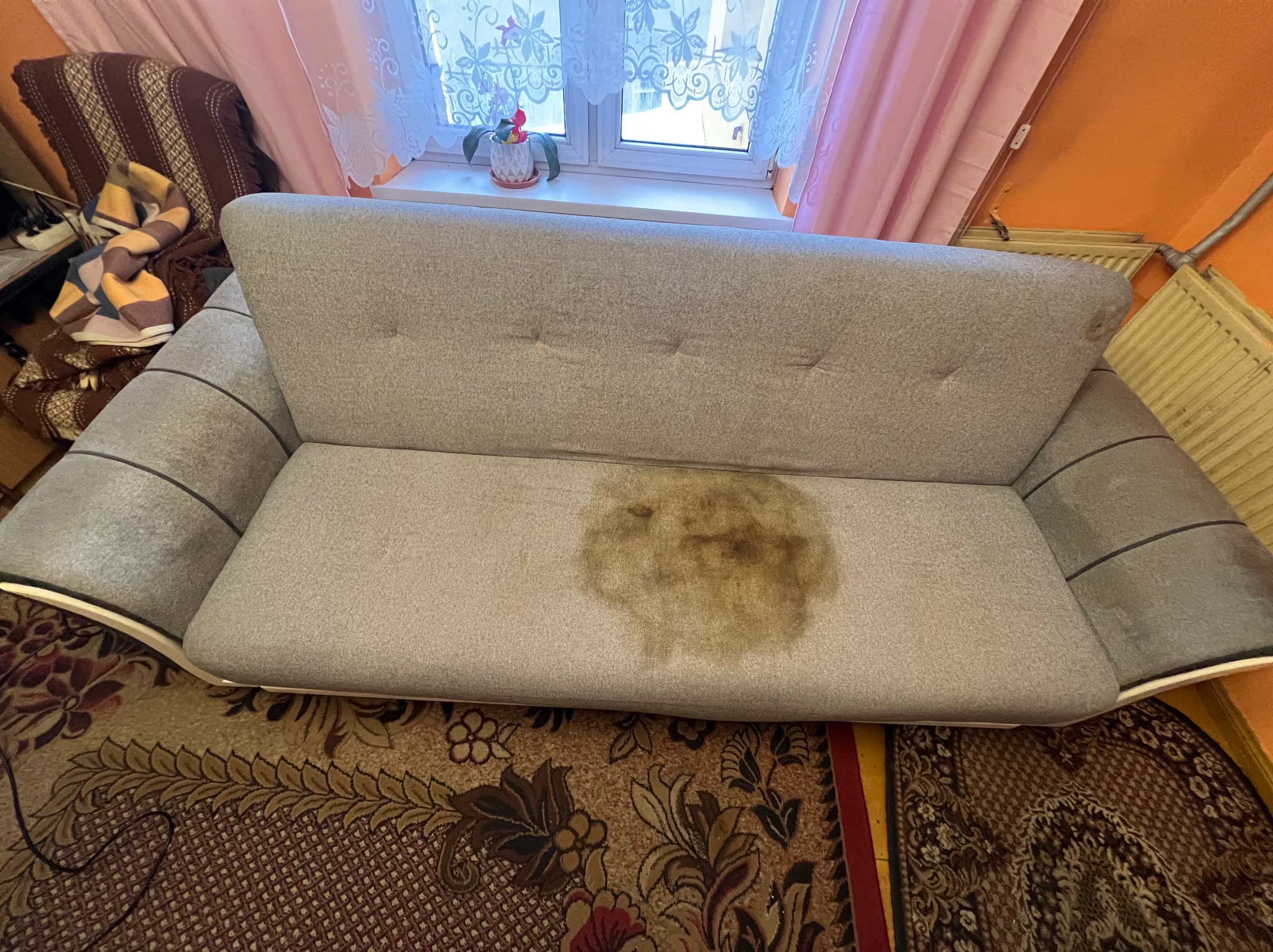 Szara kanapa z widoczną, dużą plamą na siedzisku, stojąca na wzorzystym dywanie. W tle okno z białymi firankami i różowymi zasłonami.