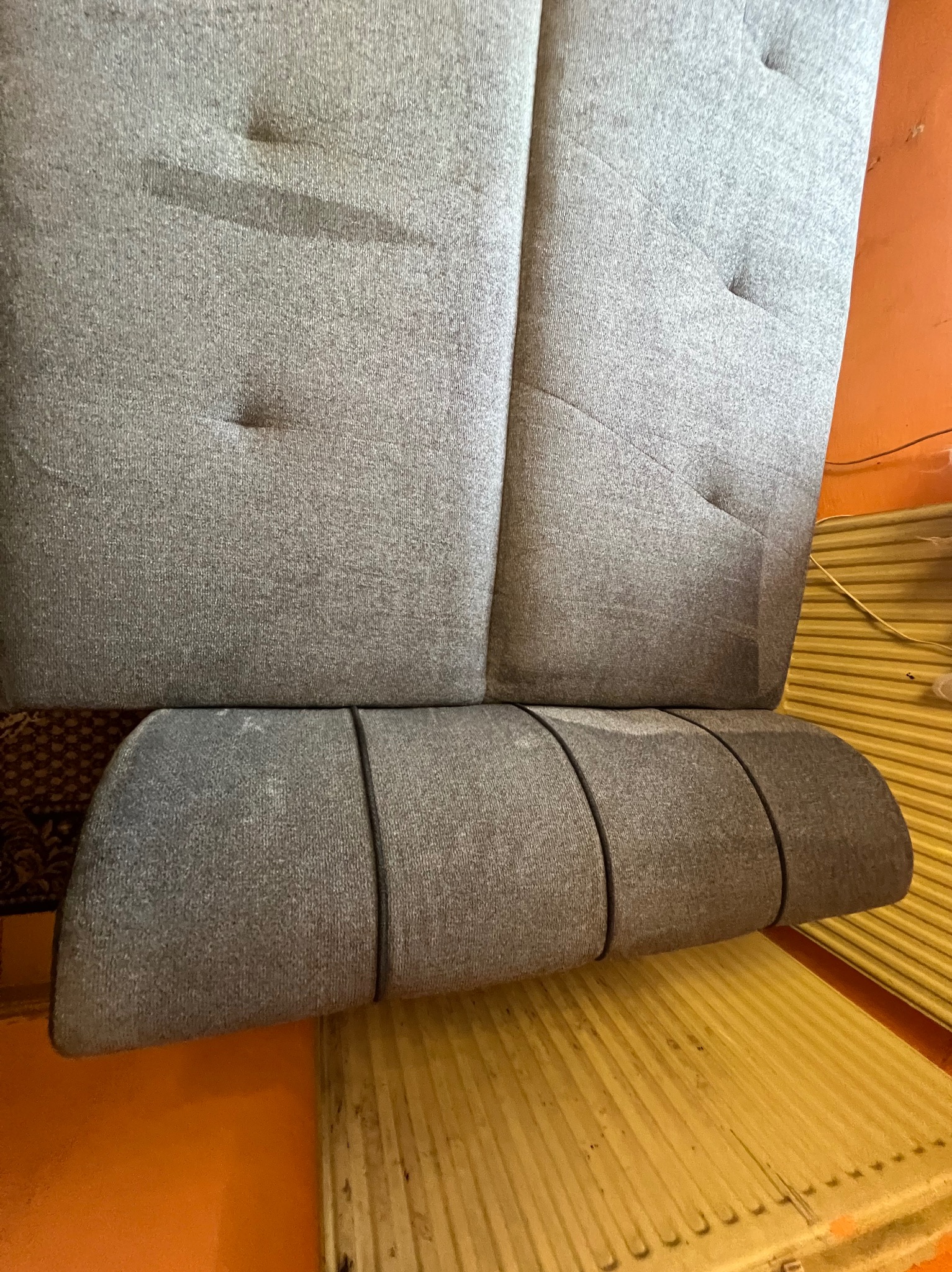 Szary zagłówek tapicerowany z guzikami, widoczne plamy i zabrudzenia na materiale. Ujęcie z bliska, ukazujące strukturę tkaniny i detale wykończenia.