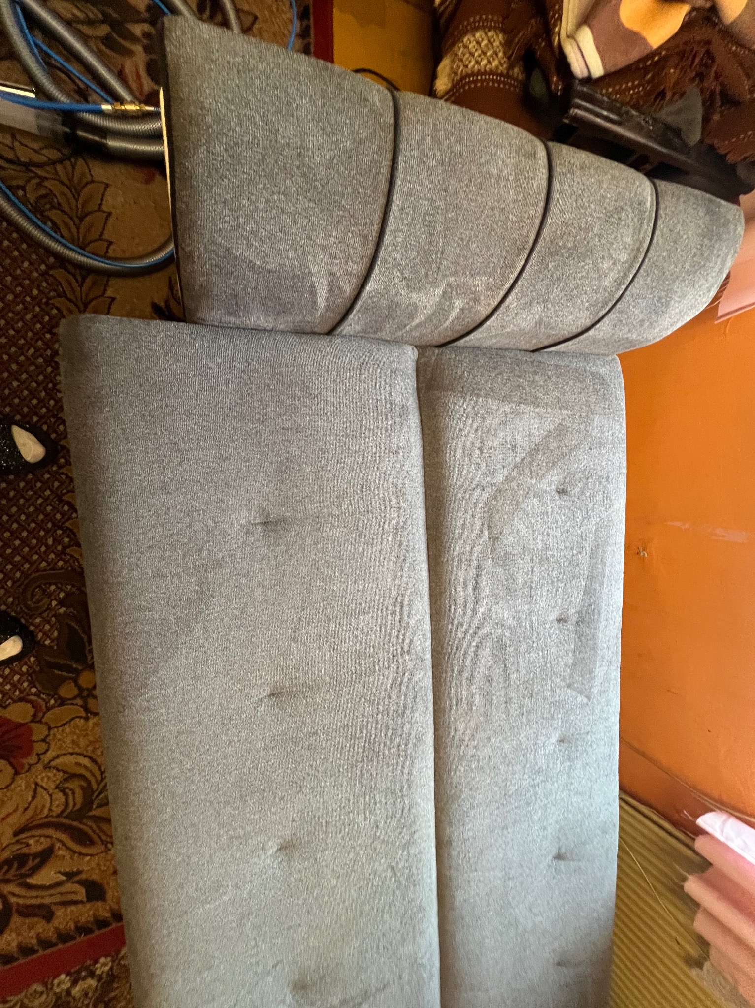 Szara kanapa z widocznymi śladami czyszczenia, fragment węża odkurzacza w tle. Widoczne różnice w odcieniu po praniu tapicerki.