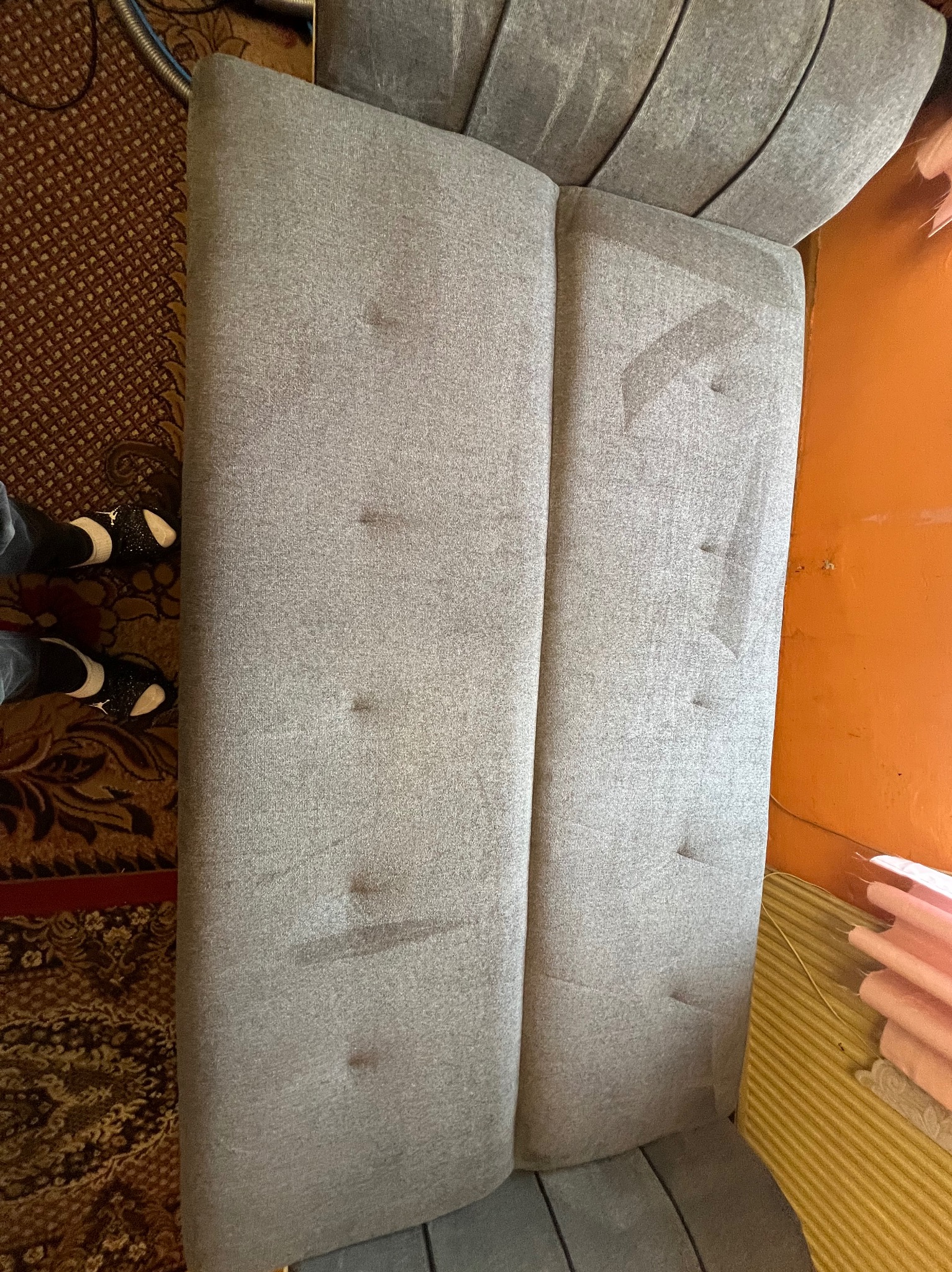 Szara kanapa z widocznymi plamami na tapicerce, sfotografowana z góry, obok dywan z wzorem i fragment pomarańczowej ściany. Widoczne buty osoby stojącej obok.