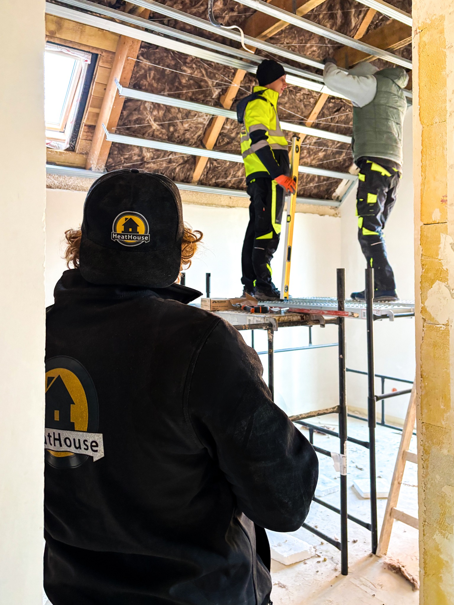 Pracownicy na rusztowaniu montują izolację poddasza z wełny mineralnej. Widoczny fragment okna dachowego i narzędzia pracy. Jeden z pracowników ubrany w odzież z logo firmy.