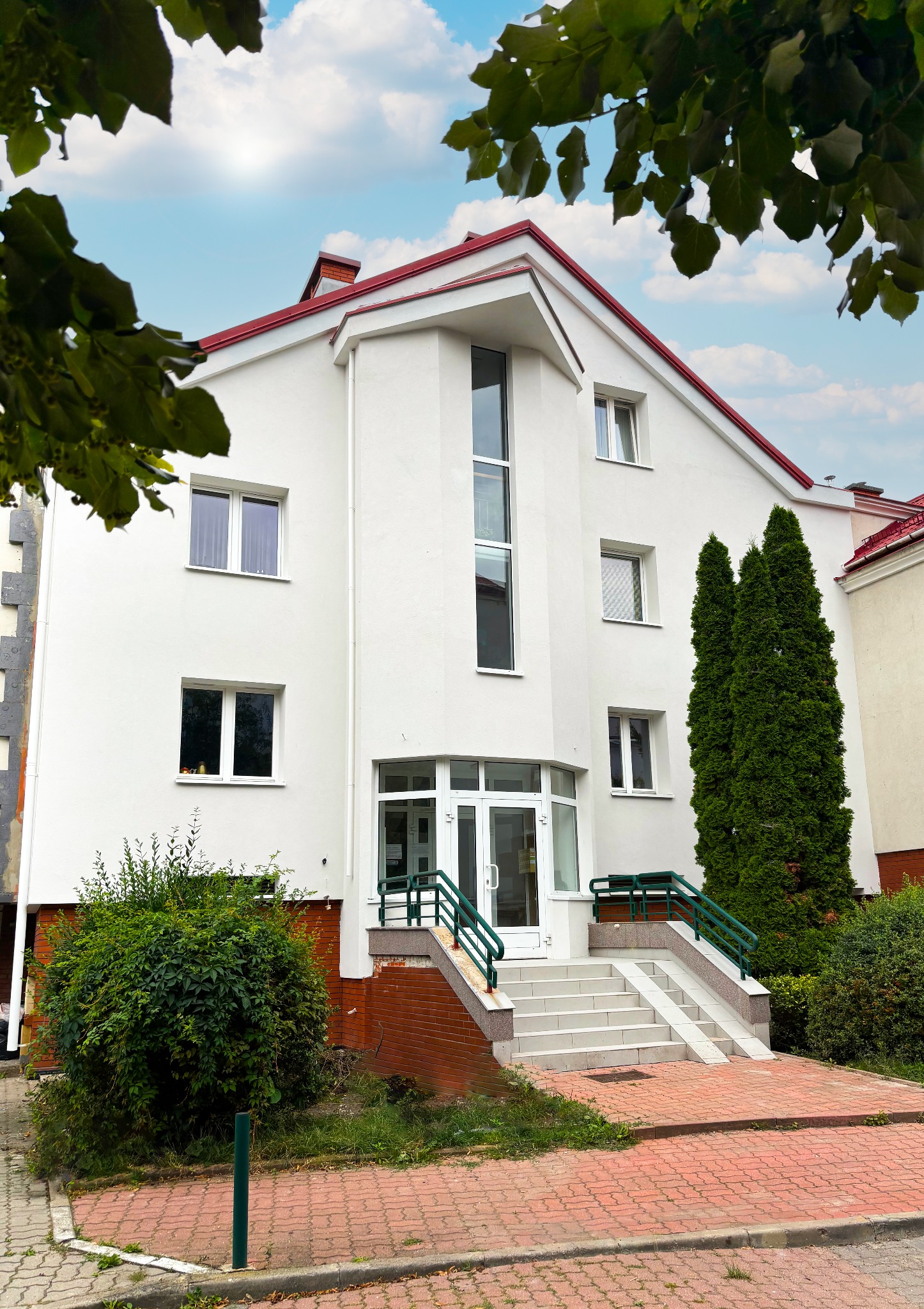 Biały budynek z czerwonym dachem, widok z dołu, z zielonymi akcentami na barierkach i roślinnością. Widoczne okna i przeszklone wejście.